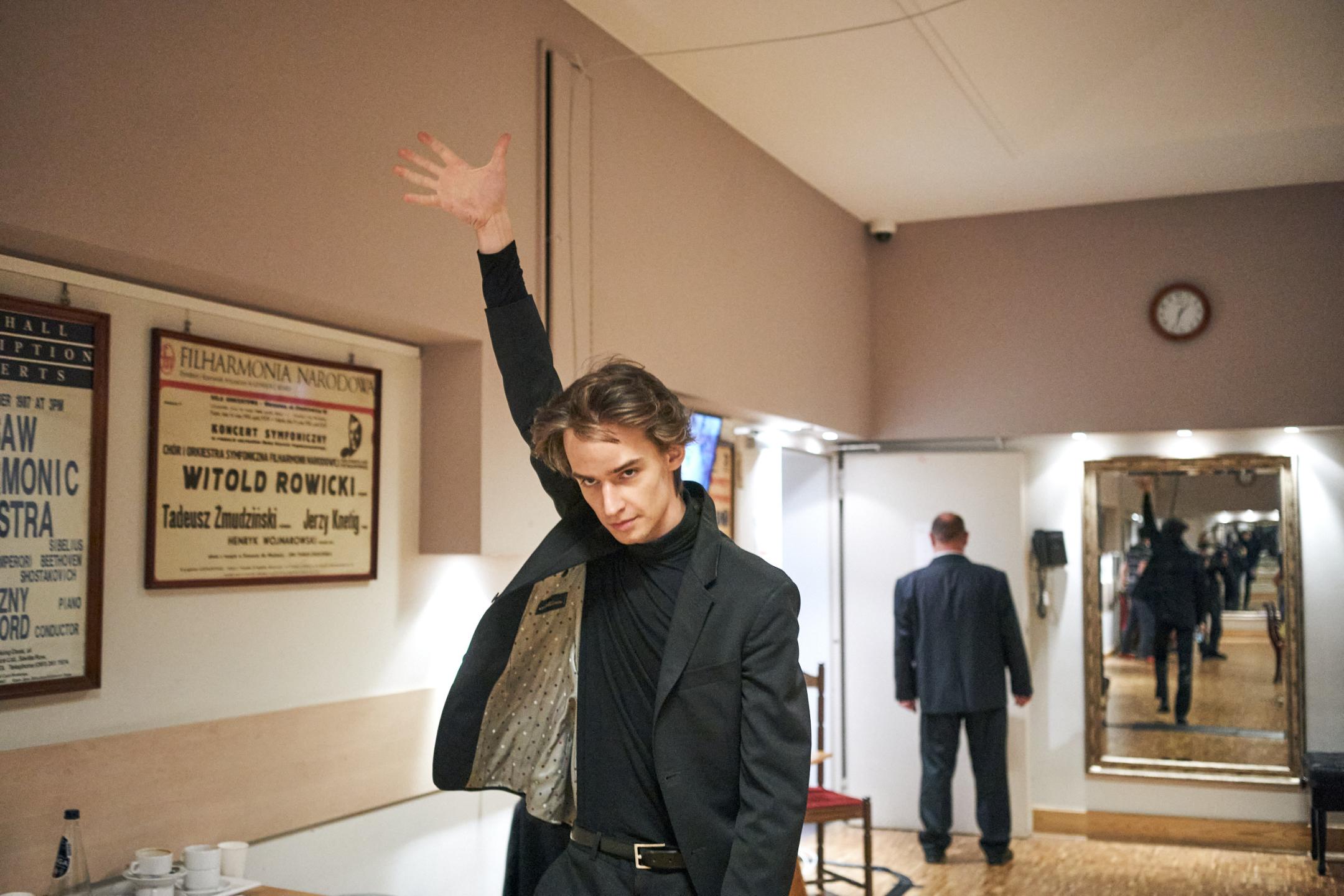 A young man in a black suit stretches his right arm and hand upwards while standing in the backstage area of a theatre.