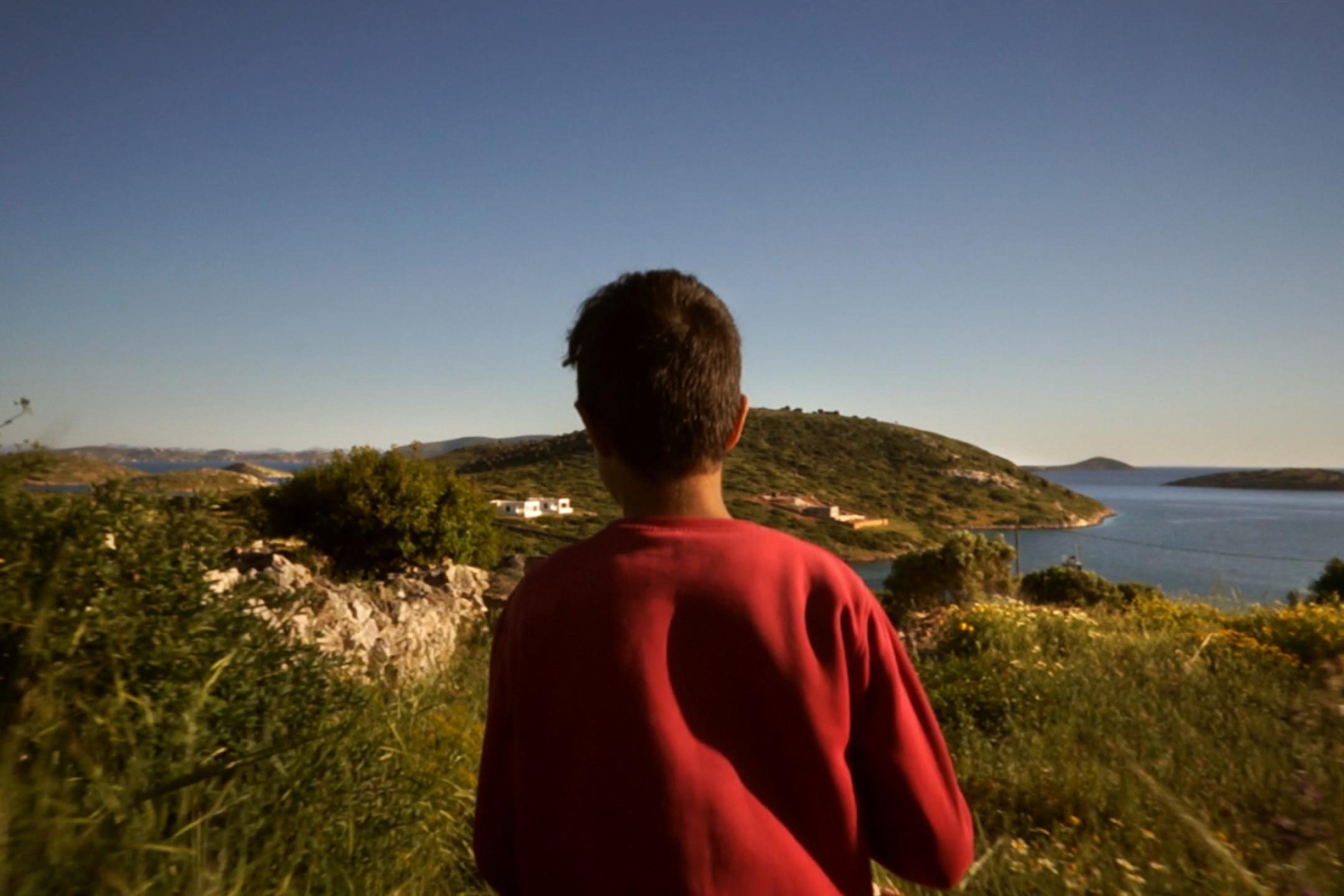A boy in a red shirt stands on top of a hill and overlooks the coastline of a small mediterranean island.