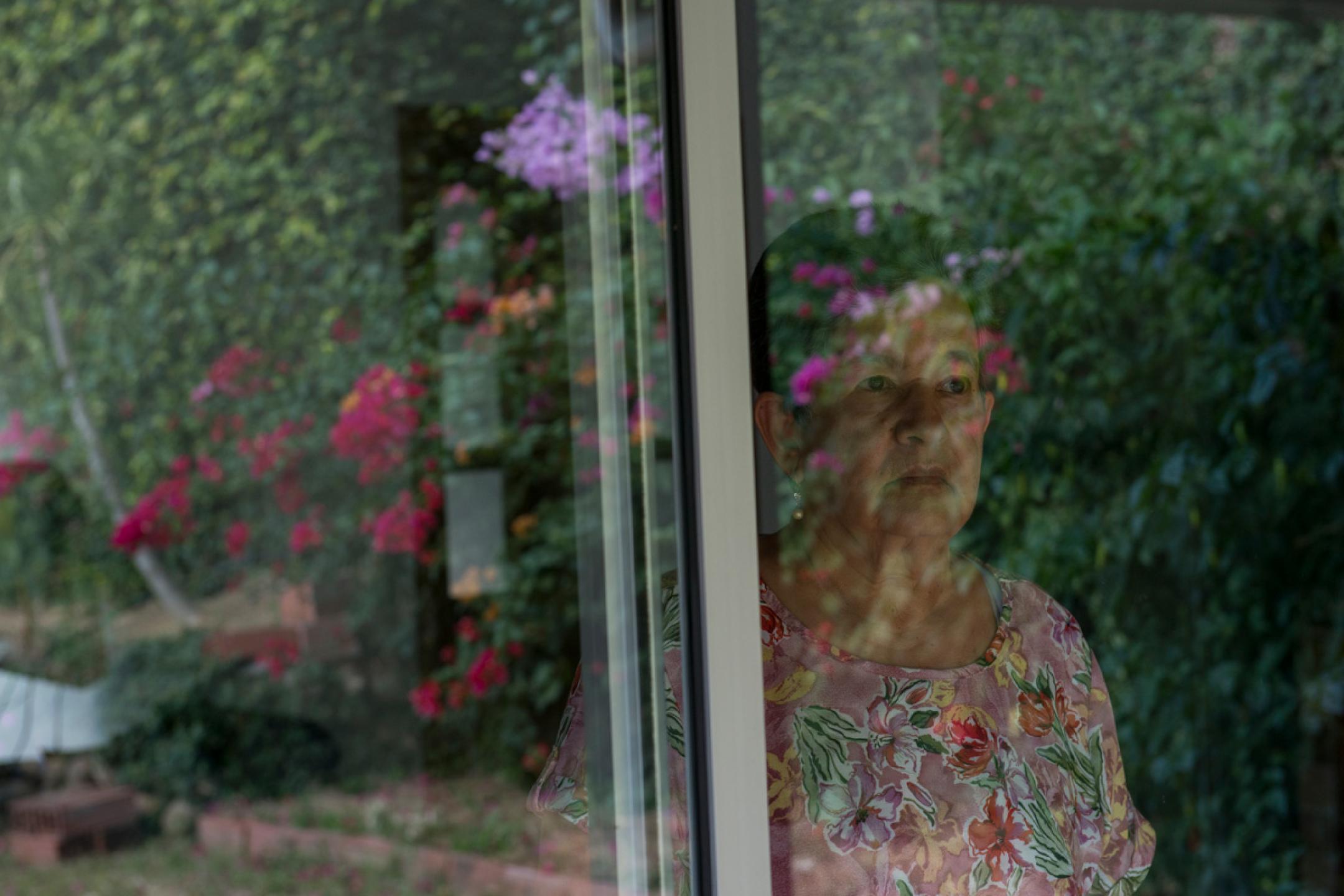 An older woman with short hair stands at a window and looks into a garden with lots of flowers.
