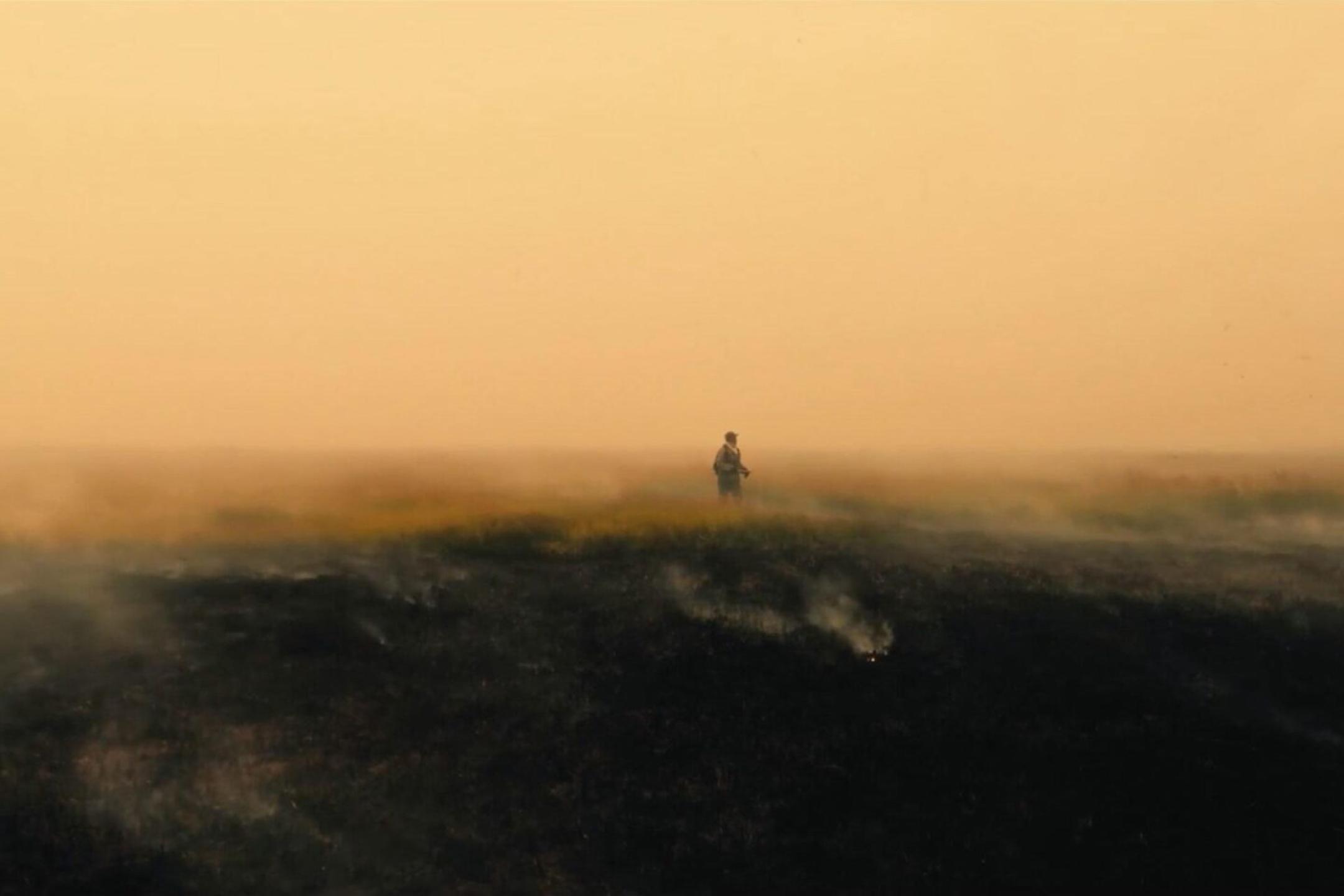 A man walks through a barren burnt landscape. Smoke rises from the ground and  clouds the view.