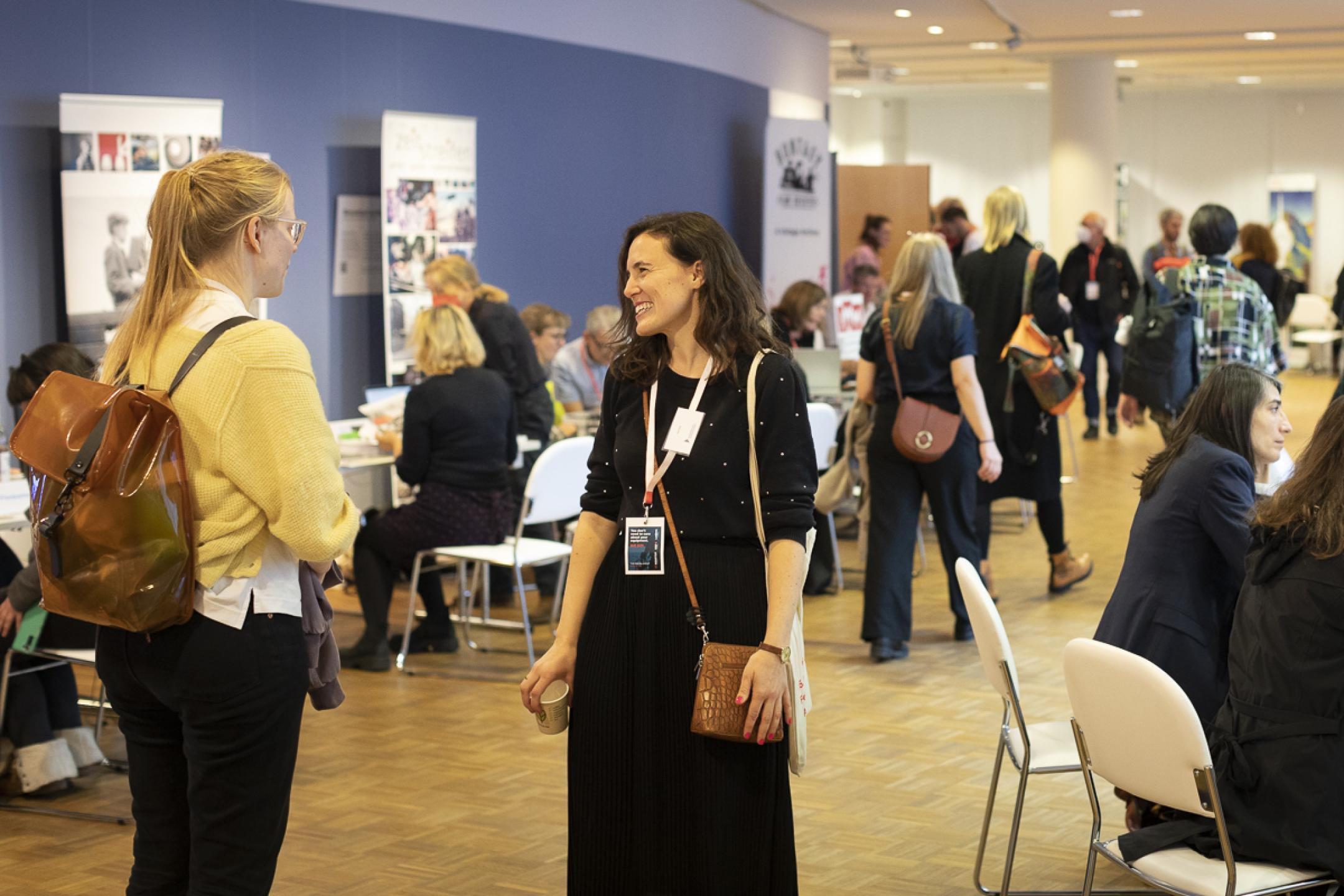 Two women are laughing and talking to each other. They are standing in the middle of a room in which meeting tables are set up at the right and left. People are sitting and standing at all the tables, talking to each other.