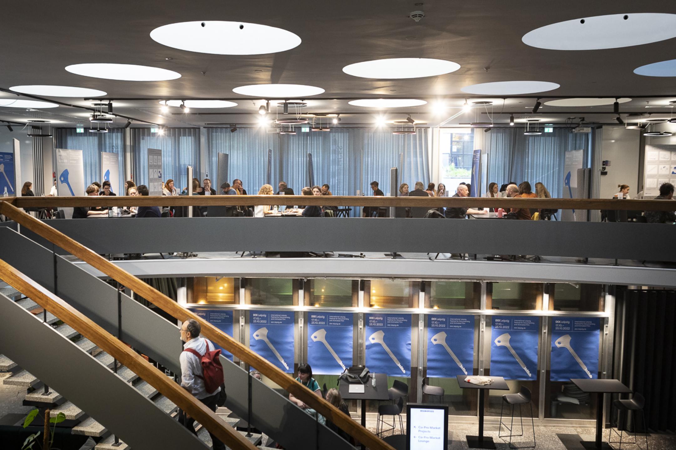 A man with a backpack goes up a staircase in an open large space in industrial  design. On the second floor there are many people sitting in small groups of three or four at tables.