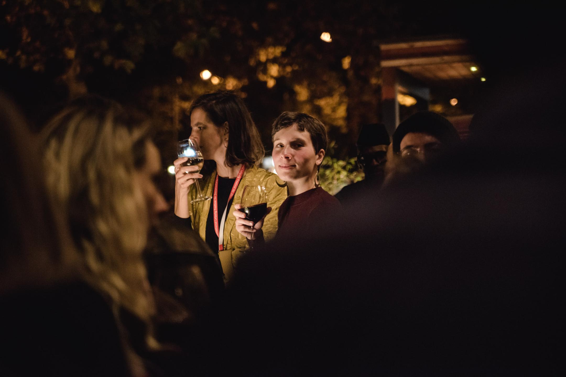Bei einer Abendveranstaltung steht eine Frau mit kurzen Haaren und einem Glas Rotwein in der Hand umringt von weiteren Personen. Sie blickt seitlich über ihre Schulter direkt in die Kamera und lächelt.