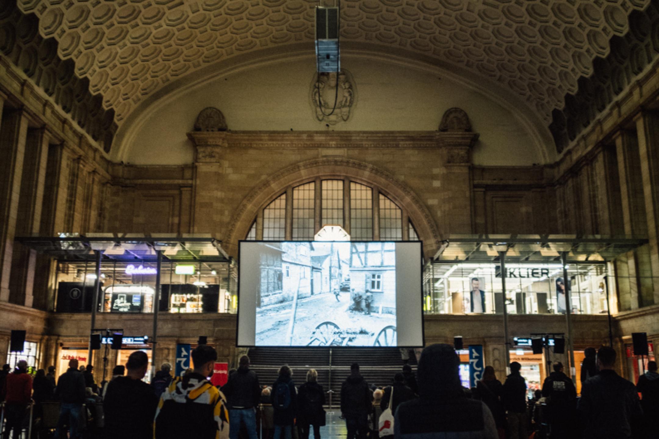 In einer großen historischen Bahnhofshalle hängt eine Kinoleinwand von der Decke, darauf ist ein schwarz-weiß Film zu sehen. Vor der Leinwand stehen und sitzen viele Personen und schauen zu.