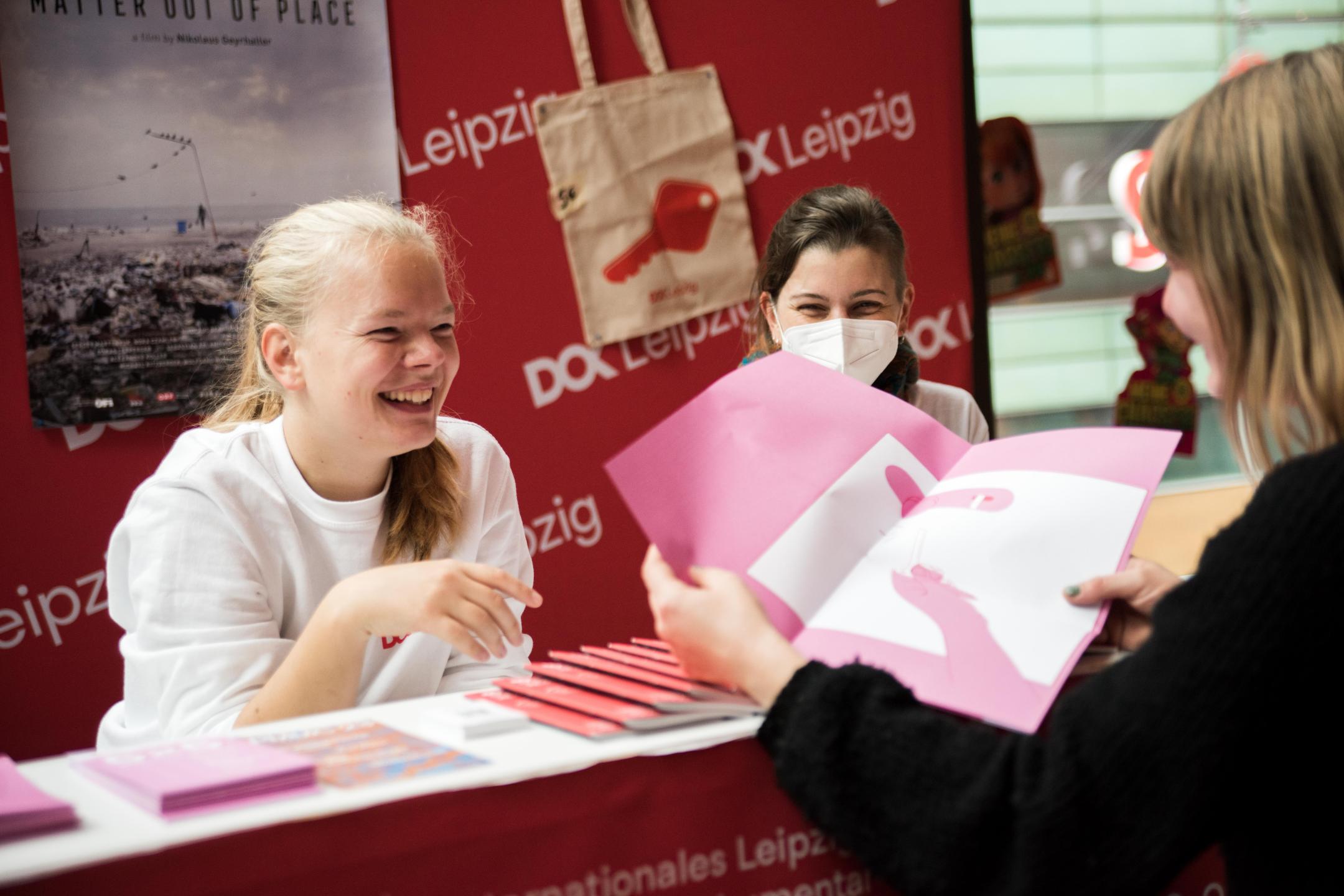 Drei Frauen stehen um einen Infostand des Festivals: Eine Besucher*in blättert in einem Flyer, zwei Frauen schauen dabei zu und lächeln.