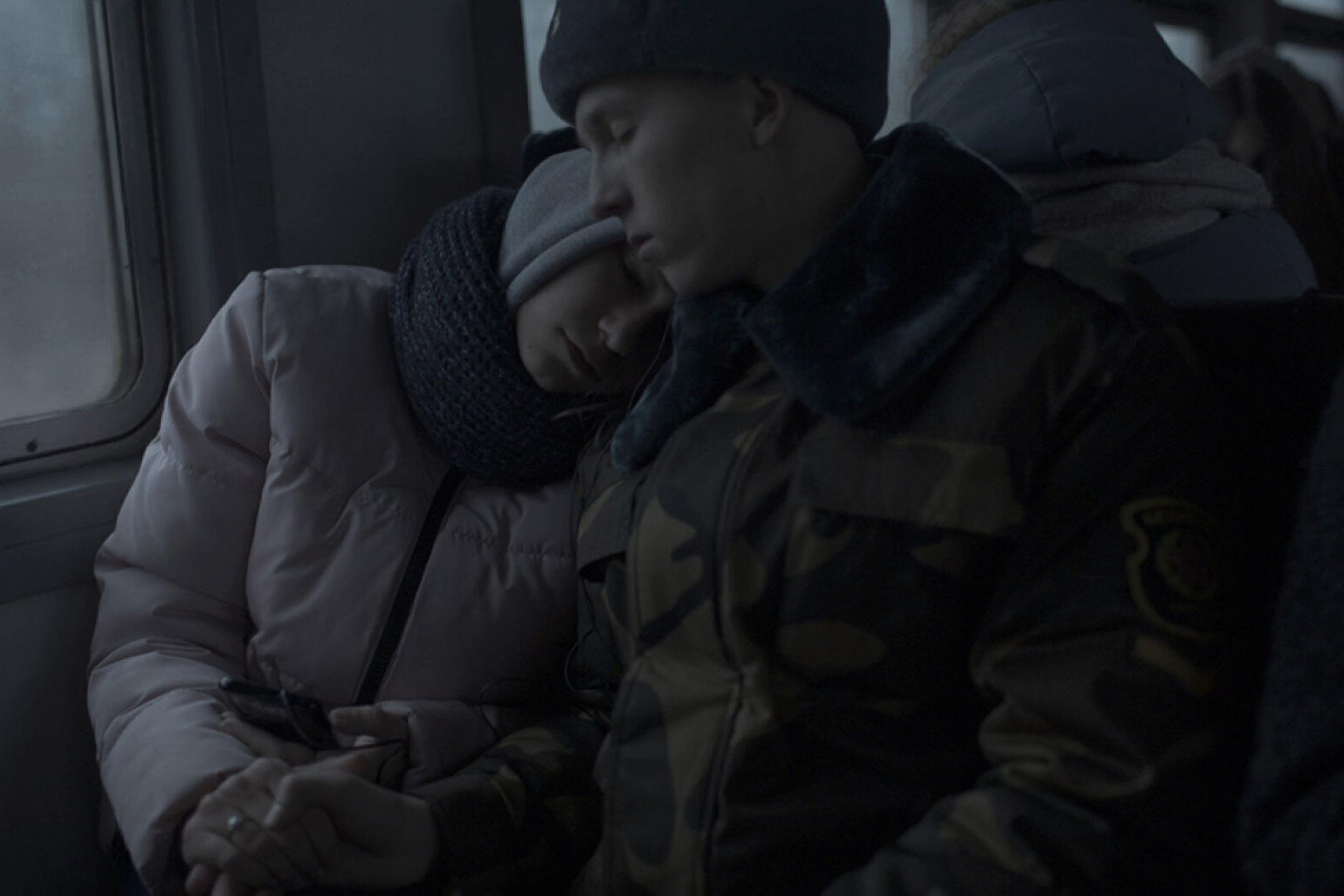 A young couple in winter clothes sits leaning at each other. Both have their eyes closed.