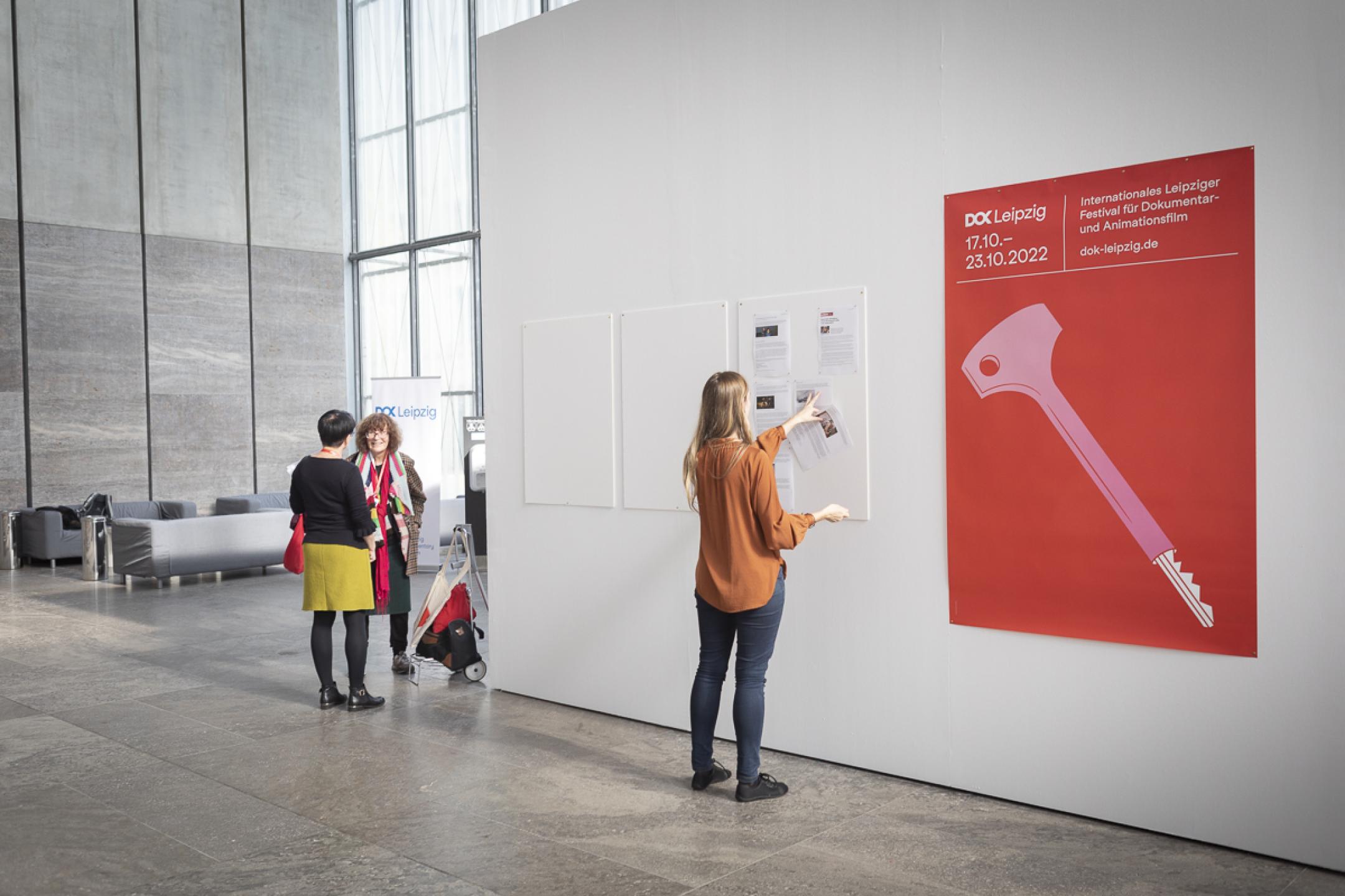 Eine Frau klebt an einer weißen hohen Wand Zeitungsausschnitte an. Daneben hängt das Festivalposter von DOK Leipzig.