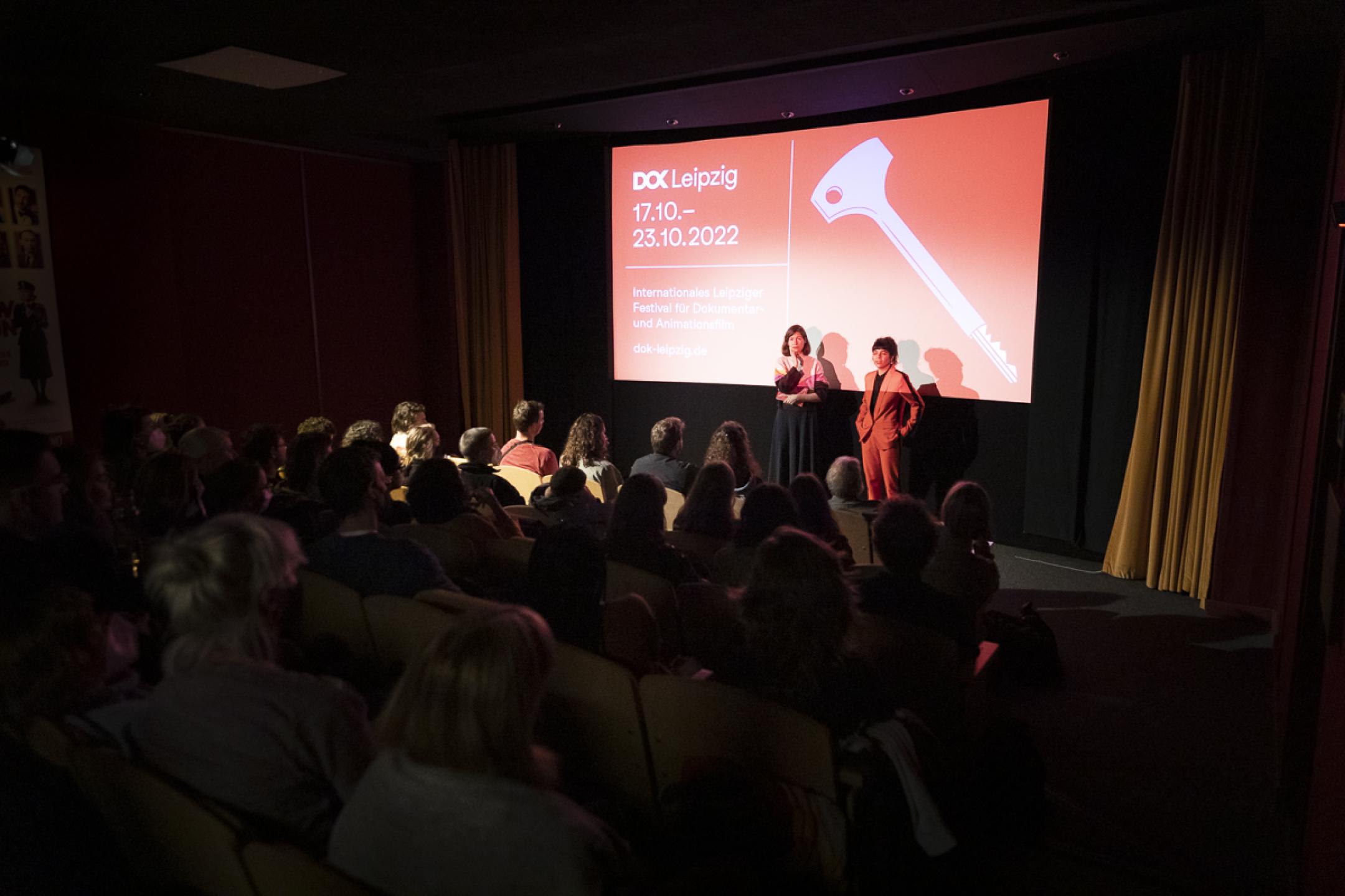 Ein vollbesetzter Kinosaal. Zwei Frauen stehen vor einer Leinwand mit dem Festivalmotiv und blicken ins Publikum.