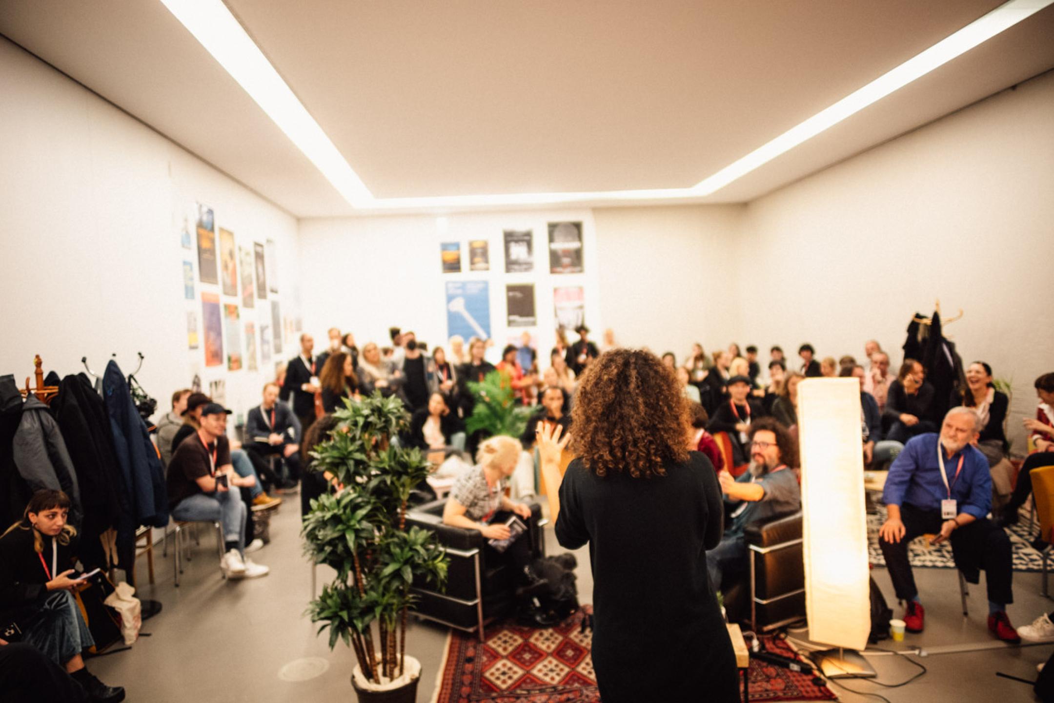 A room full of people who sit and stand while listening to a person who is speaking into a microphone.