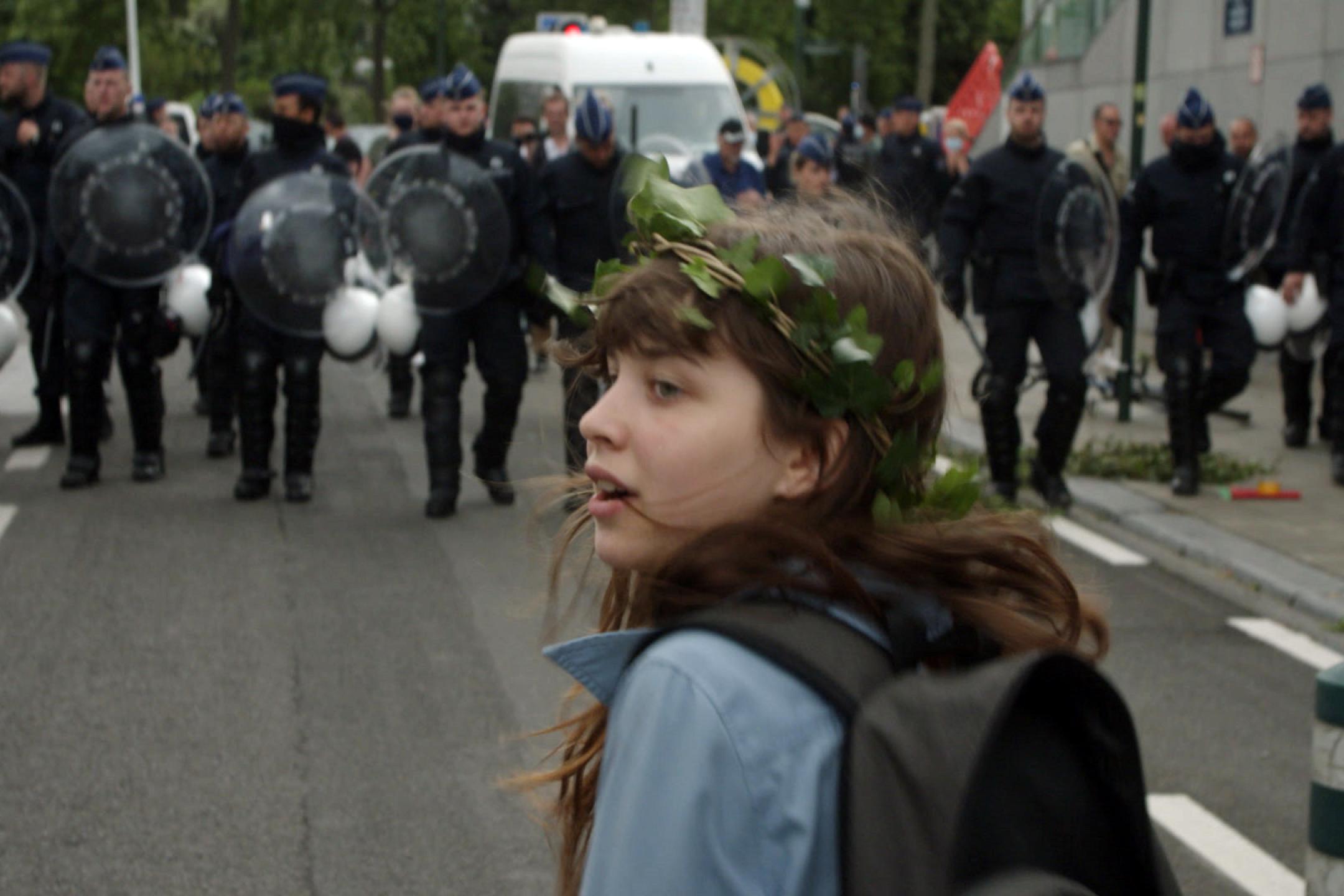 Ein Mädchen mit einem Efeu-Kranz um das lange Haar geht auf einer Straße. Ihr gegenüber stehen etwa ein Dutzend Polizisten in Uniform und mit Schutzschildern.