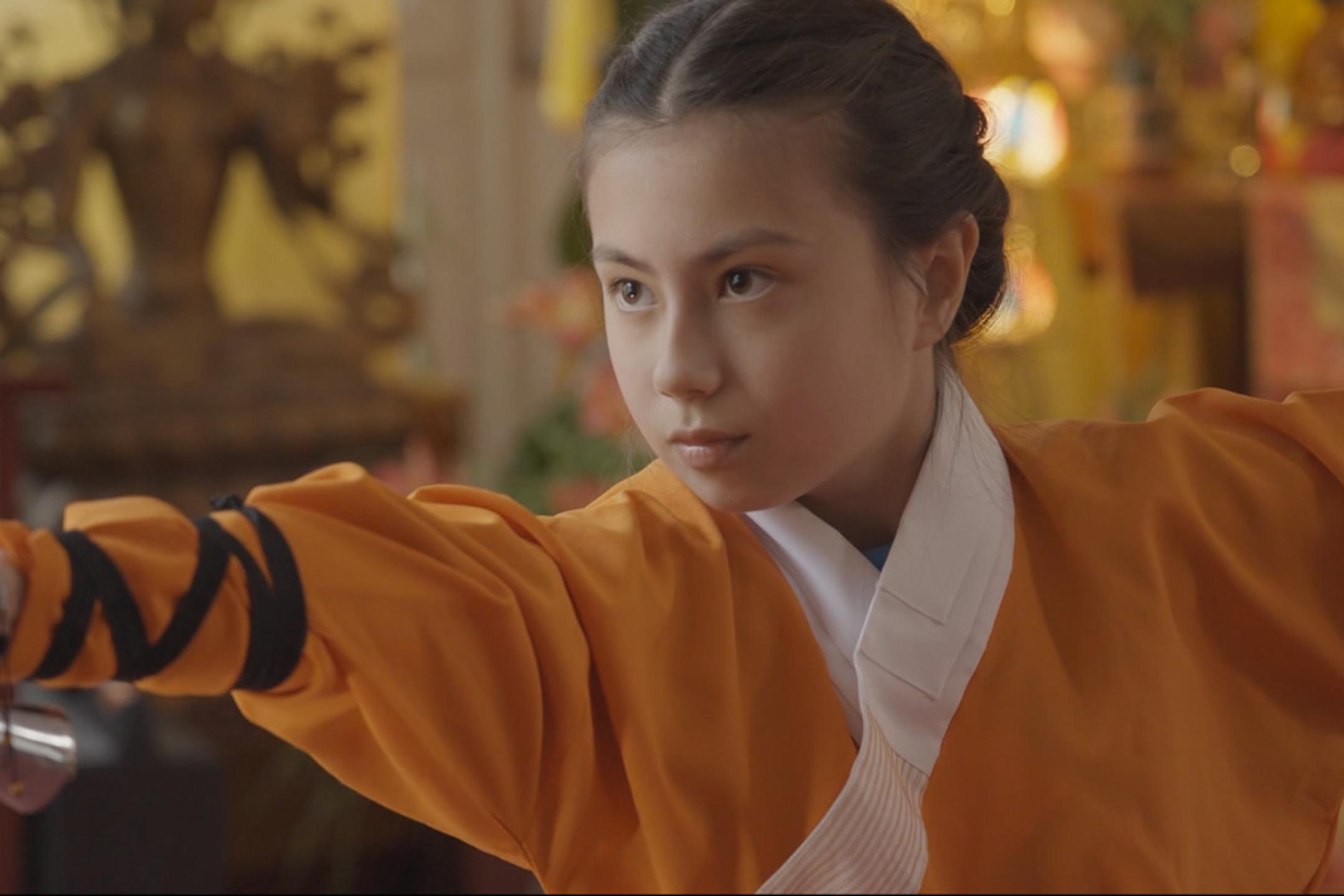 Ein Mädchen steht in einer Kung-Fu-Pose, sie trägt einen orangenen Kung-Fu-Anzug.
