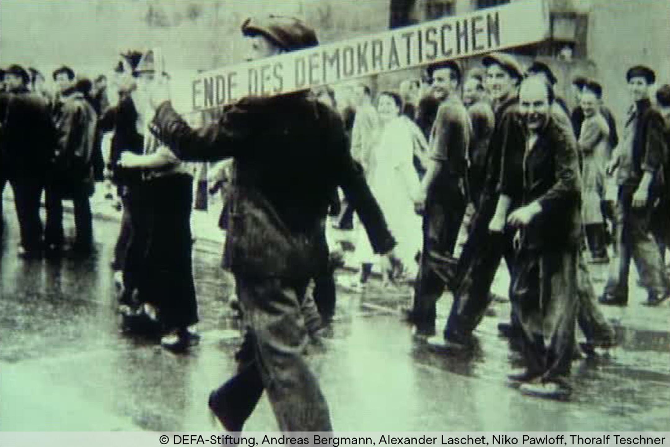 Schwarz-Weiß-Aufnahme: Mehrere Personen laufen in Gruppen auf einer Straße. Eine Person im Vordergrund trägt auf der Schulter einen langen Balken, darauf steht: "Ende des Demokratischen".