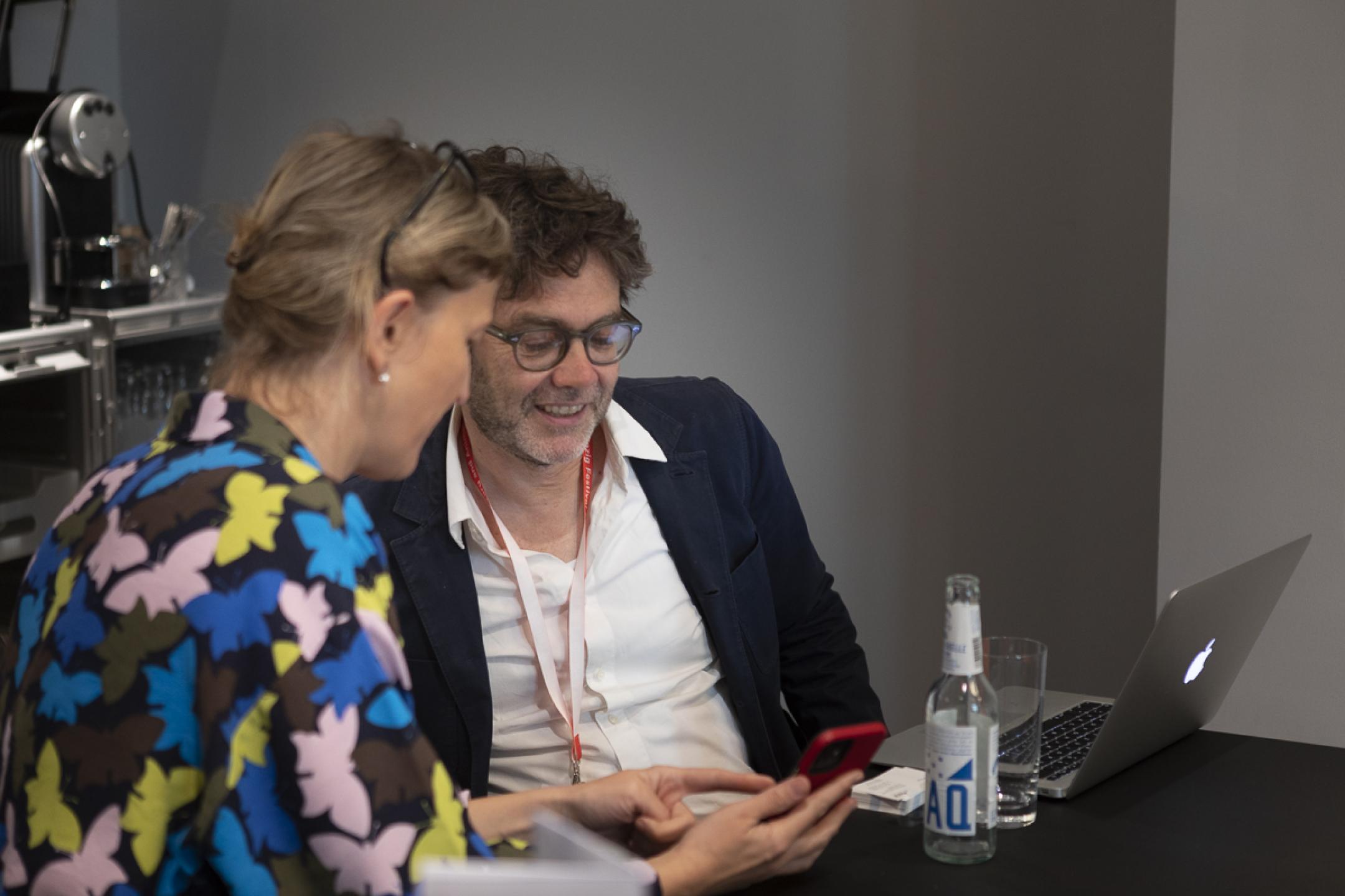 Two persons sit next to each other at a working table, looking together at one smartphone while talking. Both are smiling.