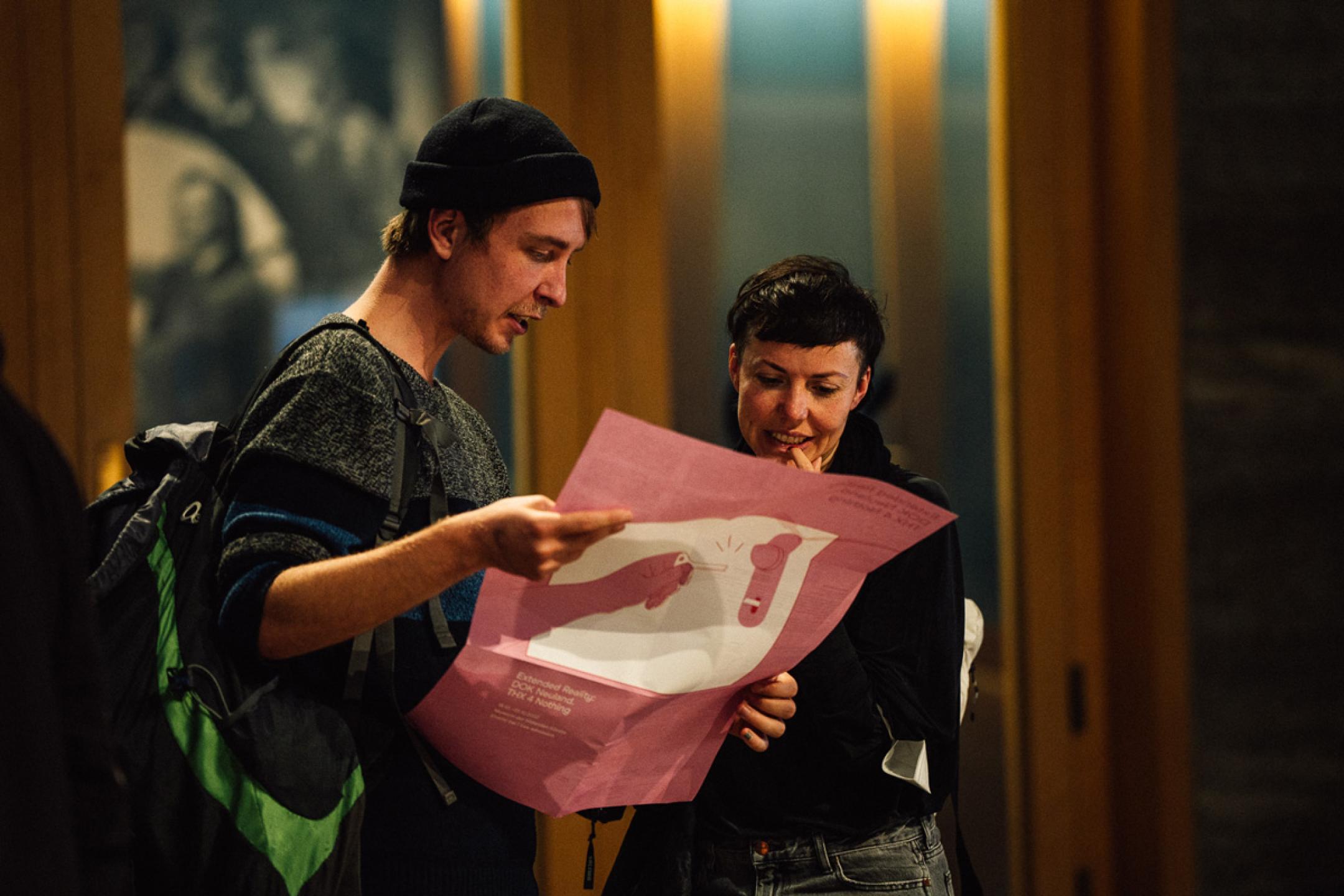 Two persons stand together and look at a DOK Leipzig flyer that the person at the left is holding in his hands.