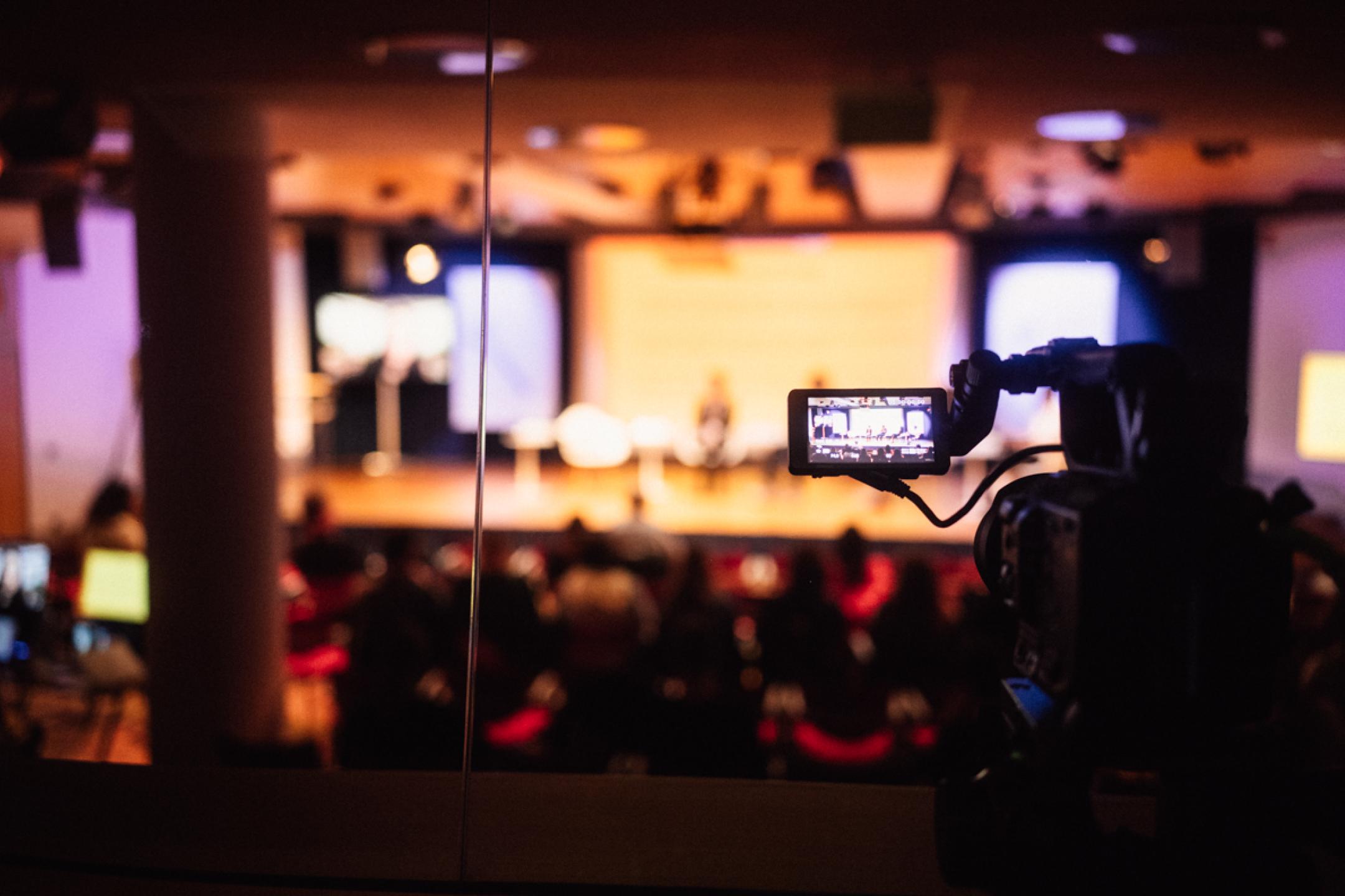 A camera monitor shows a panel of four persons on the podium. 
