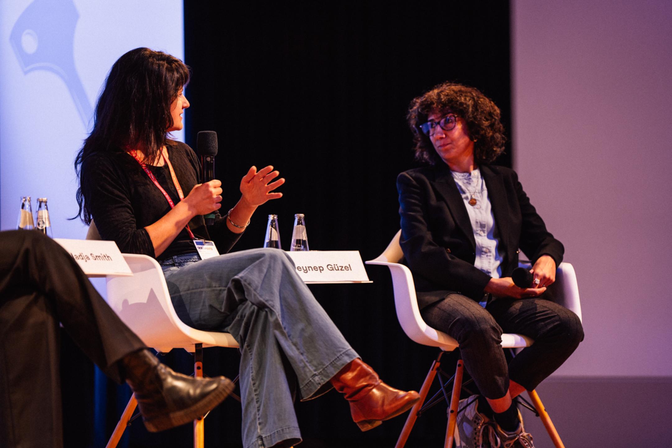 Two women sit next to each other on a panel. The person at the left speaks in a microphone.