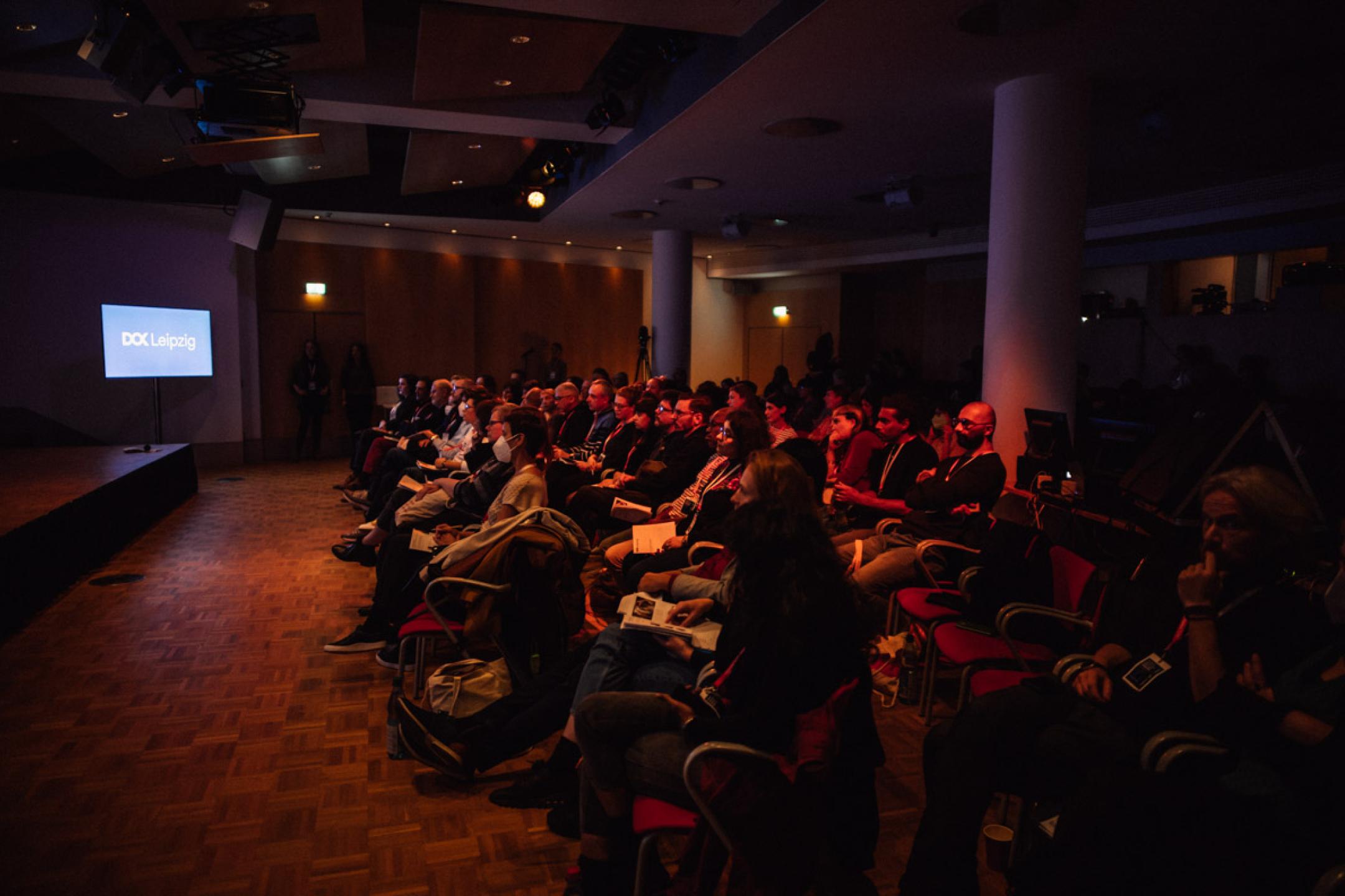 Audience in a conference room. A screen at the side shows the DOK Leipzig logo.