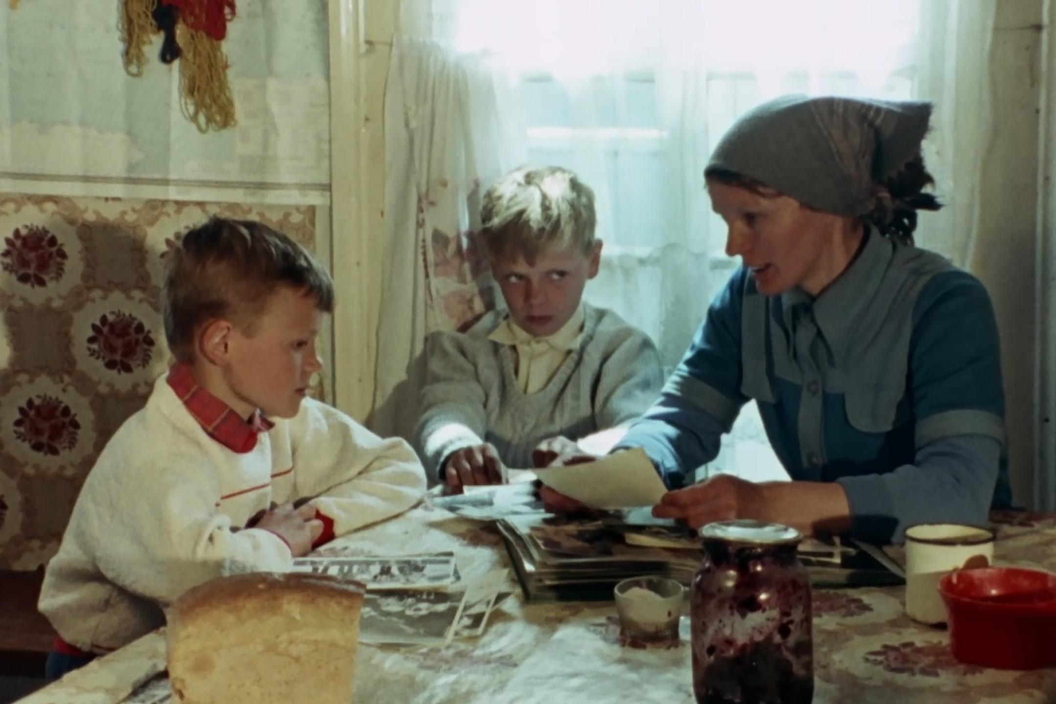 Eine Frau mit Kopftuch sitzt mit zwei Jungen an einem Tisch. Sie hält Fotos in der Hand.