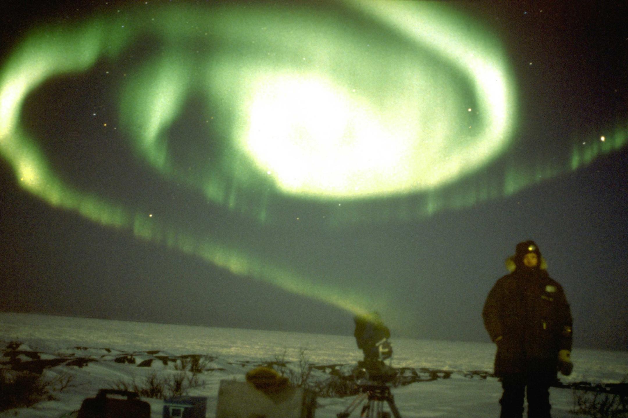 Ein Mann steht bei Nacht auf einer schneebedeckten Fläche. Über ihm strahlt ein Polarlicht Kreiselform.