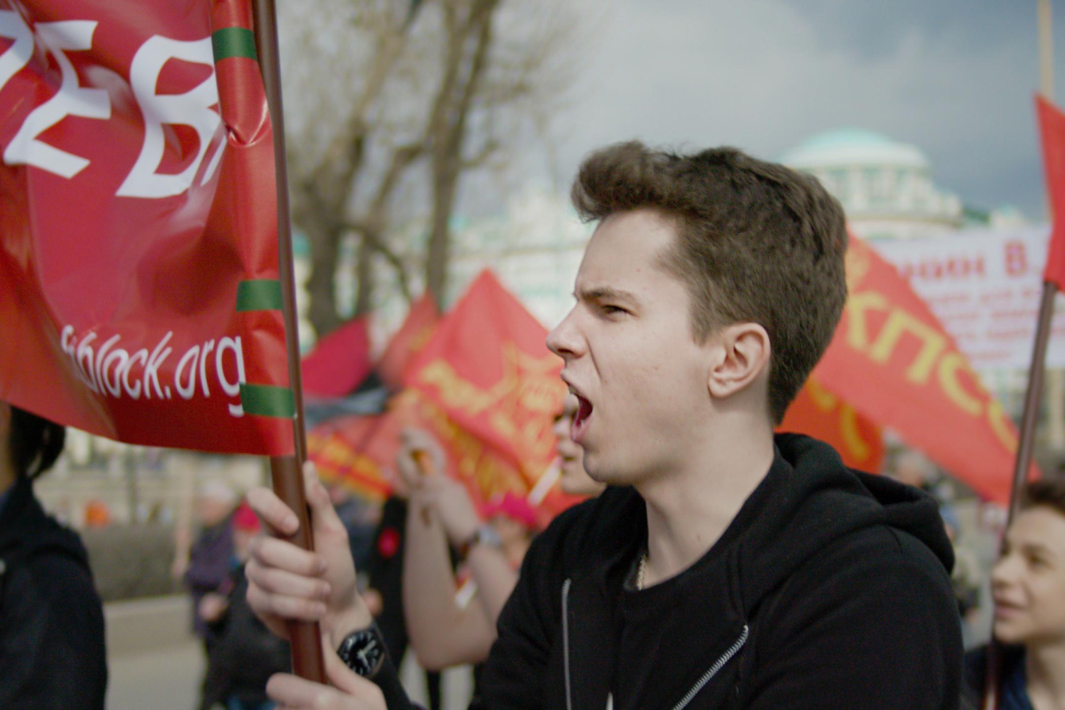 Ein junger Mann geht in einem Demonstrationszug. Er ruft etwas und hält ein großes Banner in der Hand.