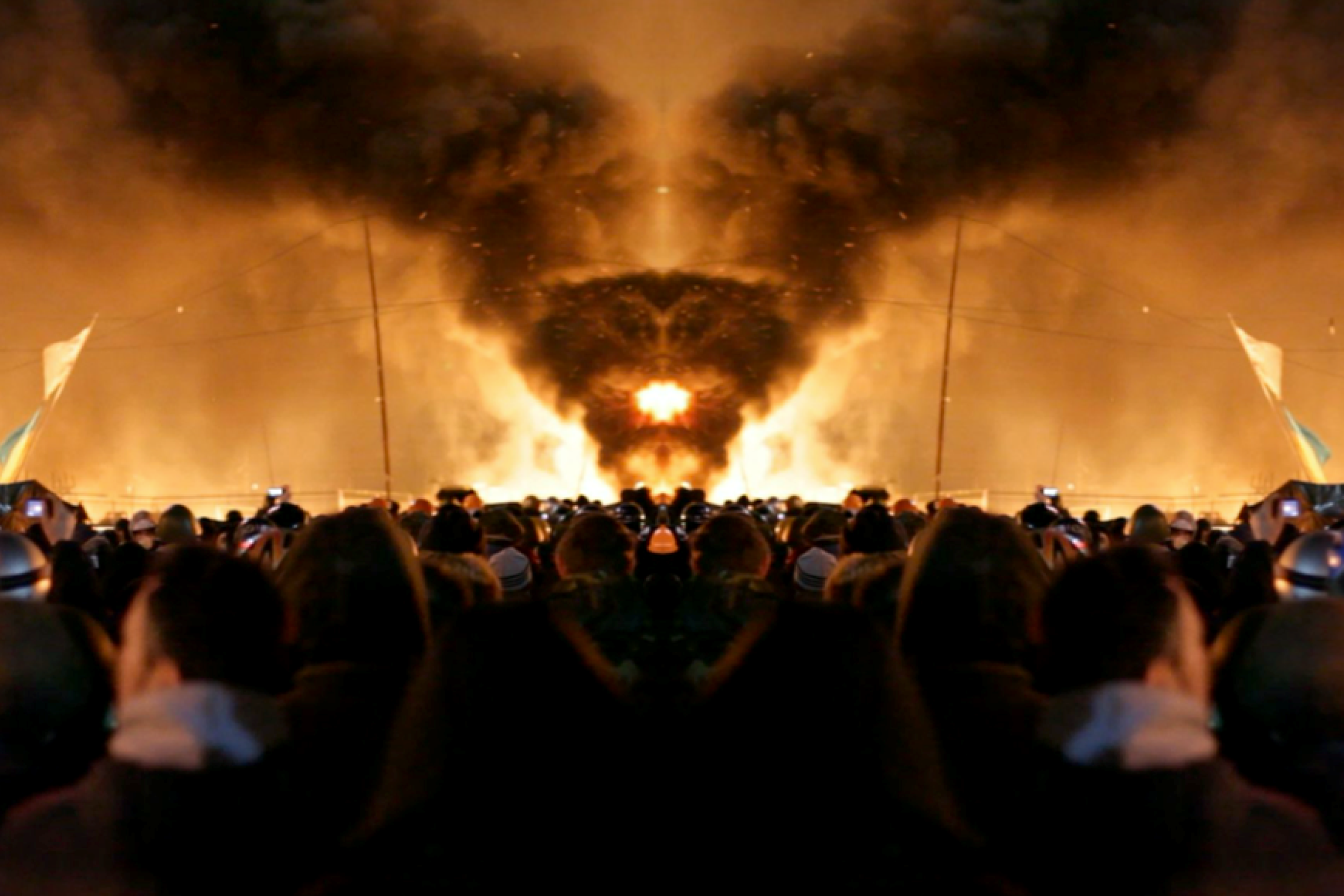 A large group of people stands close together and looks towards a column of smoke that rises in the sky.