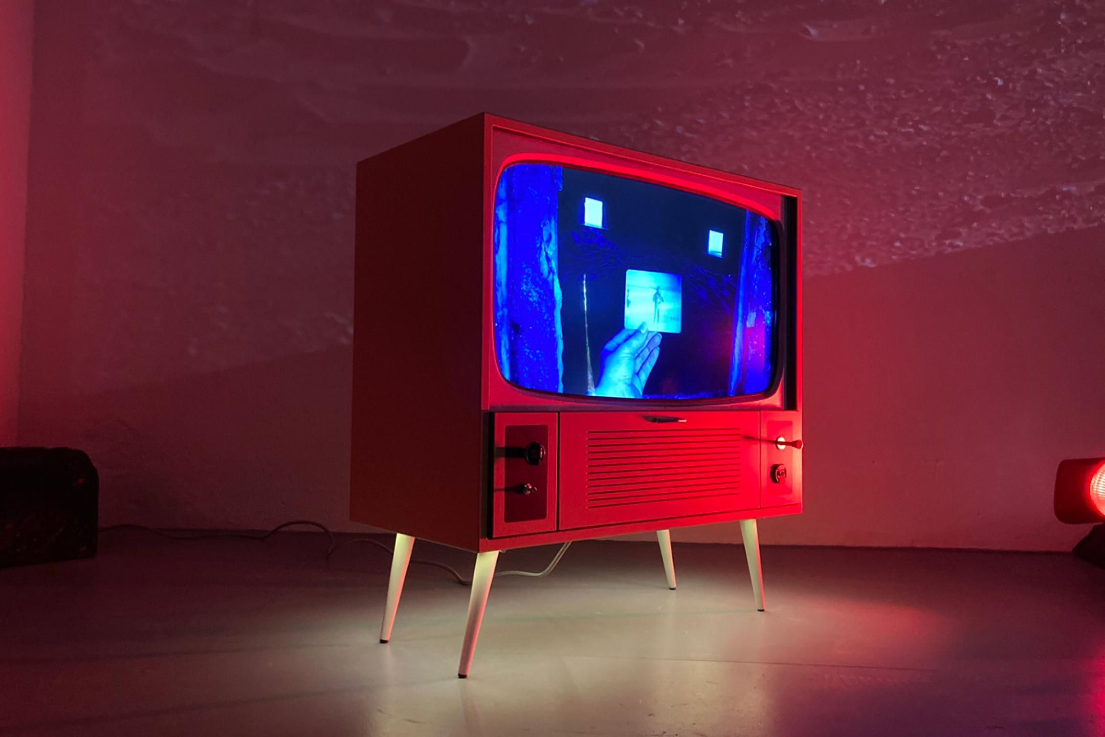 A retro TV stands in a room. On screen you can a hand that holds a photo of a person at the beach.
