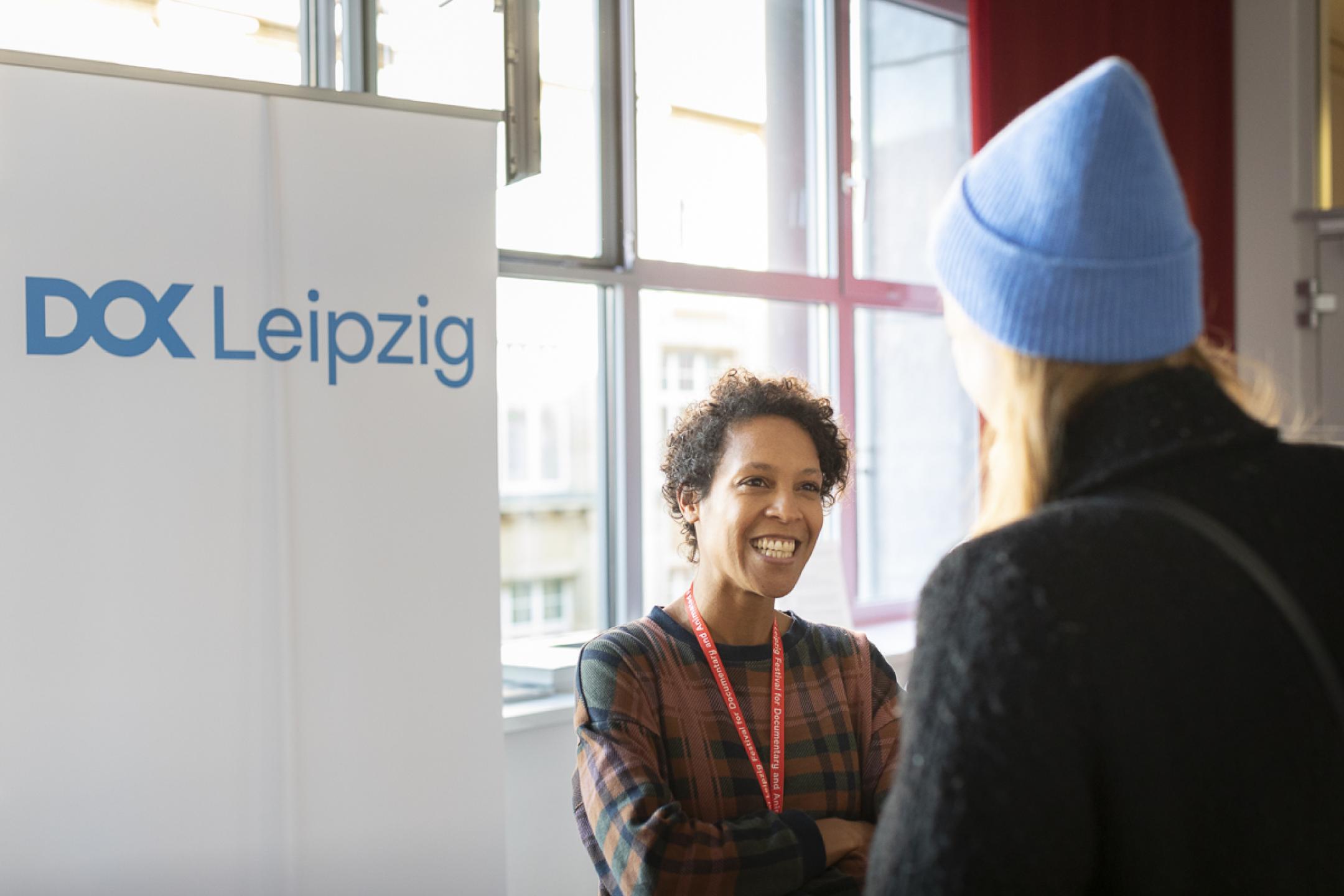 Two persons stand in front of a DOK Leipzig banner and talk.