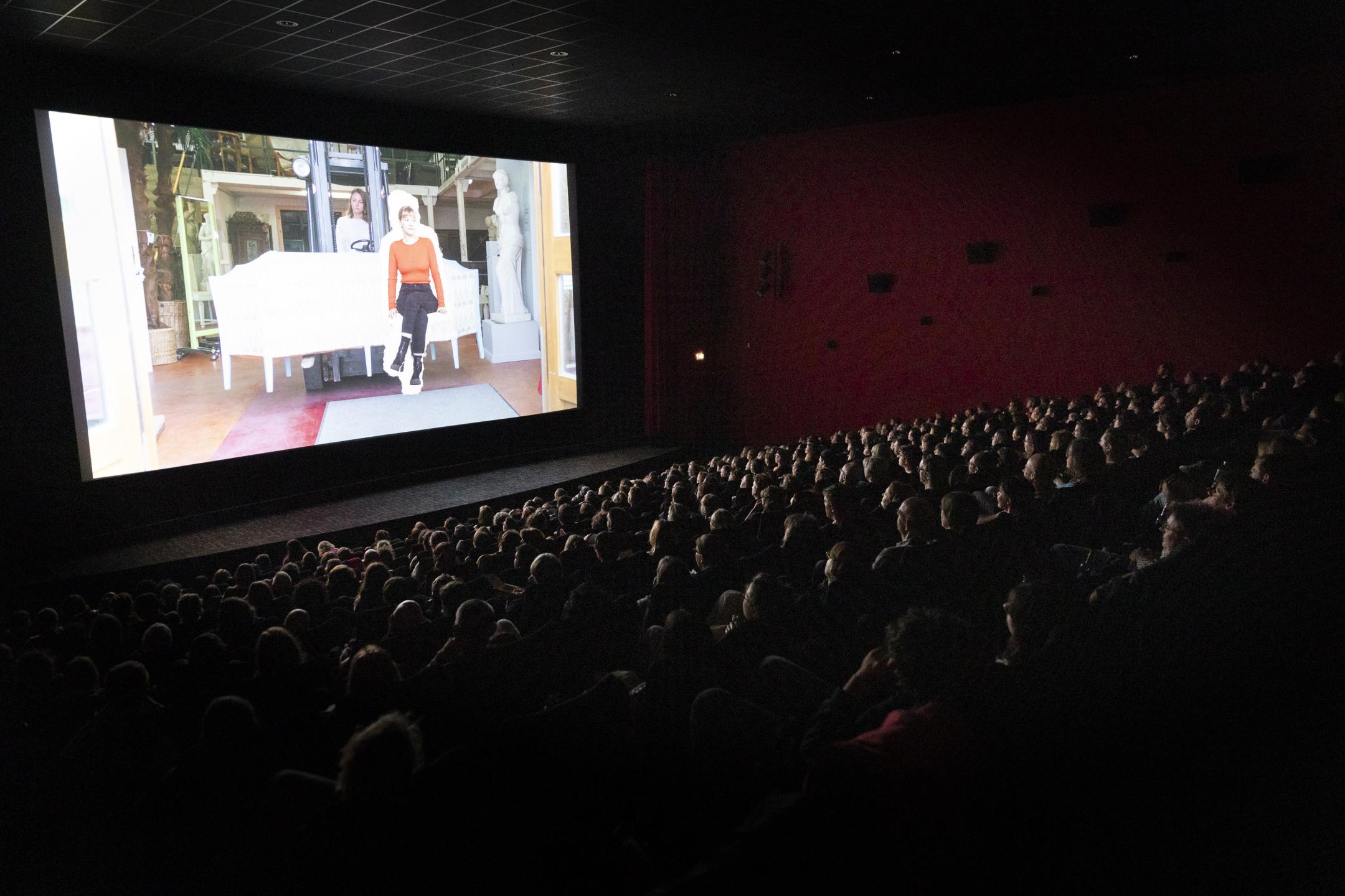 Ein voll besetzter Kinosaal bei einer Filmvorführung. Auf der Leinwand eine Frau, die auf einem Sofa sitzt.