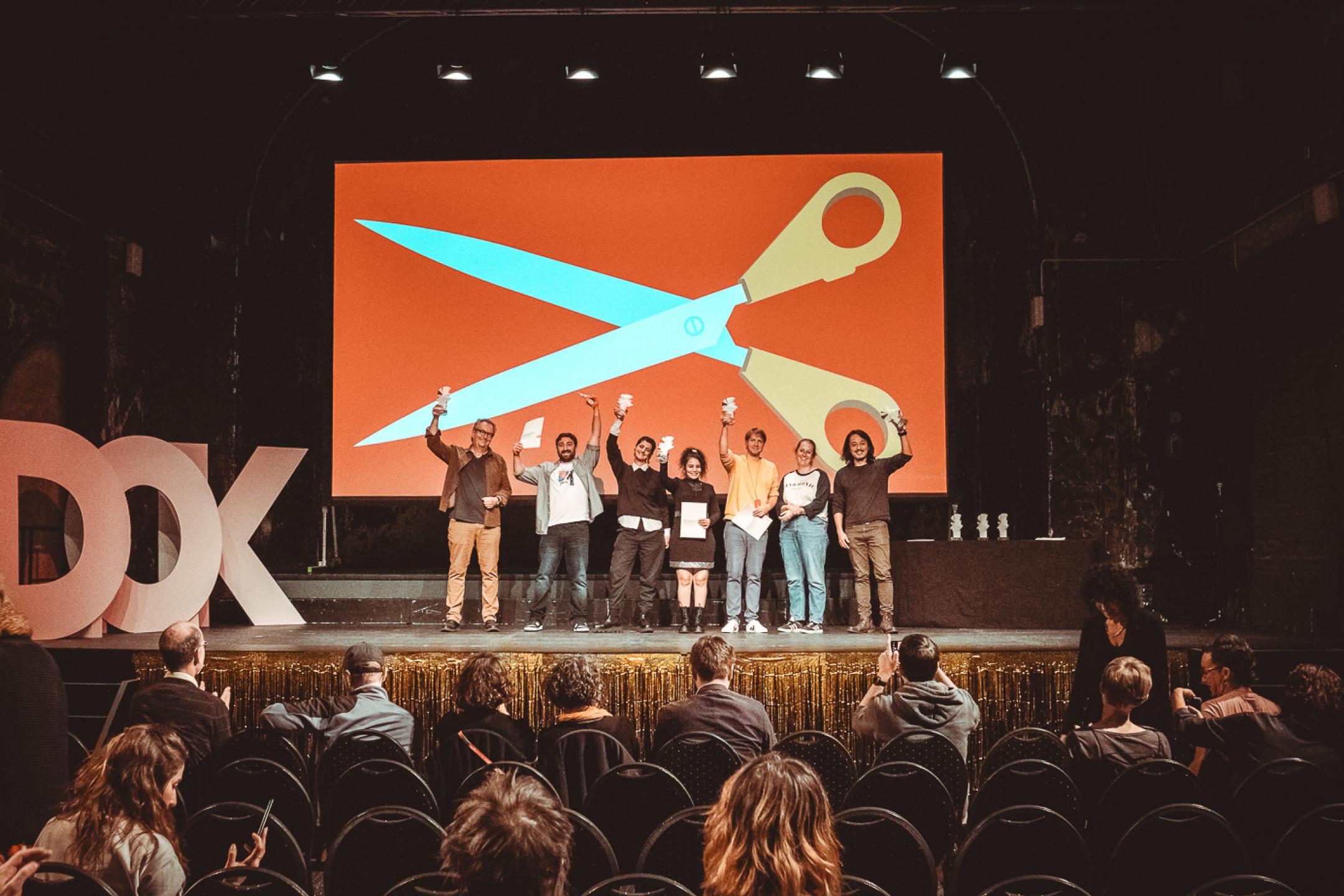 Sieben Personen stehen auf einer Bühne und halten Preisstatuetten in die Höhe. Im Hintergrund das Festivalmotiv von DOK Leipzig 2023: eine riesige Schere.