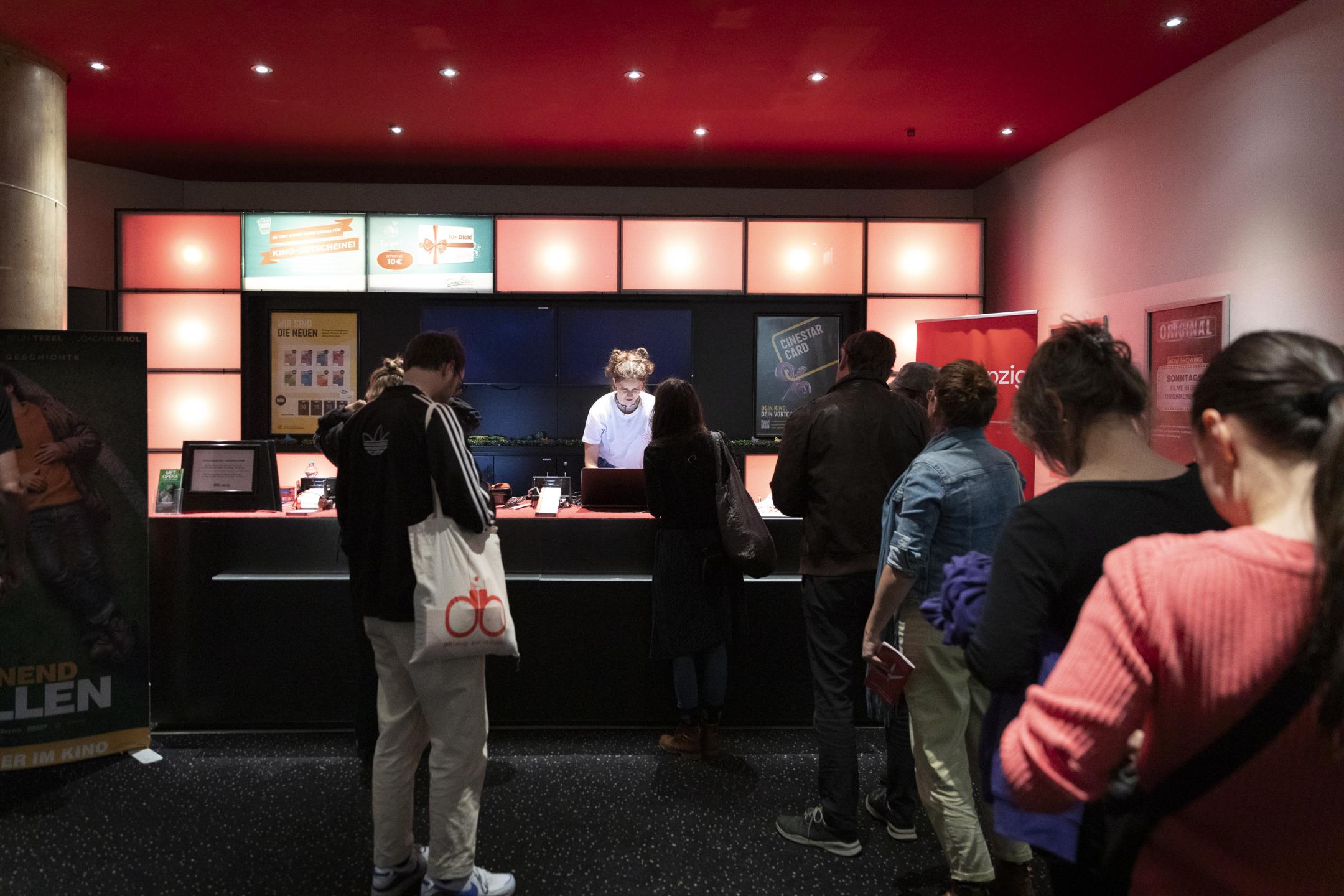 Mehrere Personen stehen an einer Kinokasse in der Schlange. Eine Mitarbeiterin in einem weißen T-Shirt steht hinter dem Kassentresen.