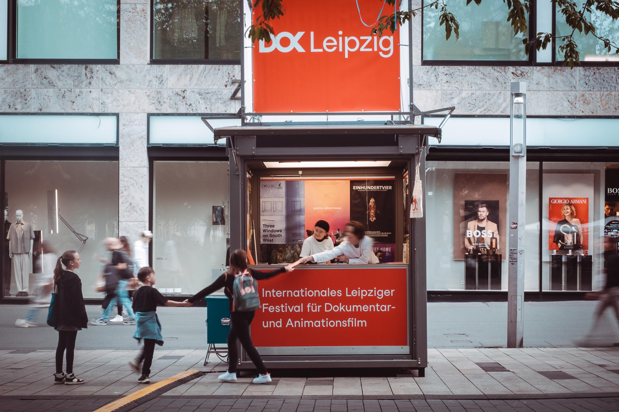 Zwei Personen in einer Info-Box in einer Fußgängerzone. Eine der Personen reicht einen Flyer an vorbeigehende Kinder.
