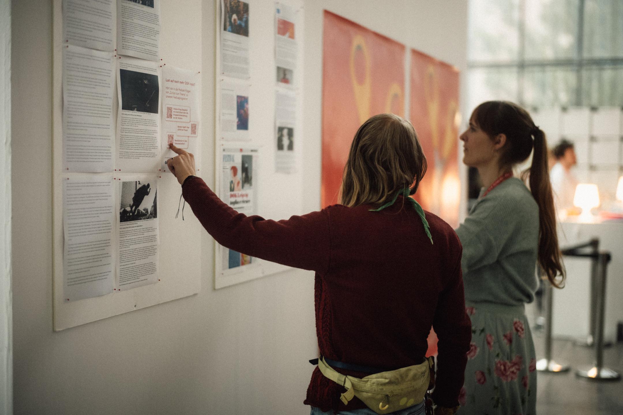 Zwei Frauen stehen an einer Wand mit Aushängen und Festivalplakaten. Eine Frau zeigt auf einen der Aushänge.