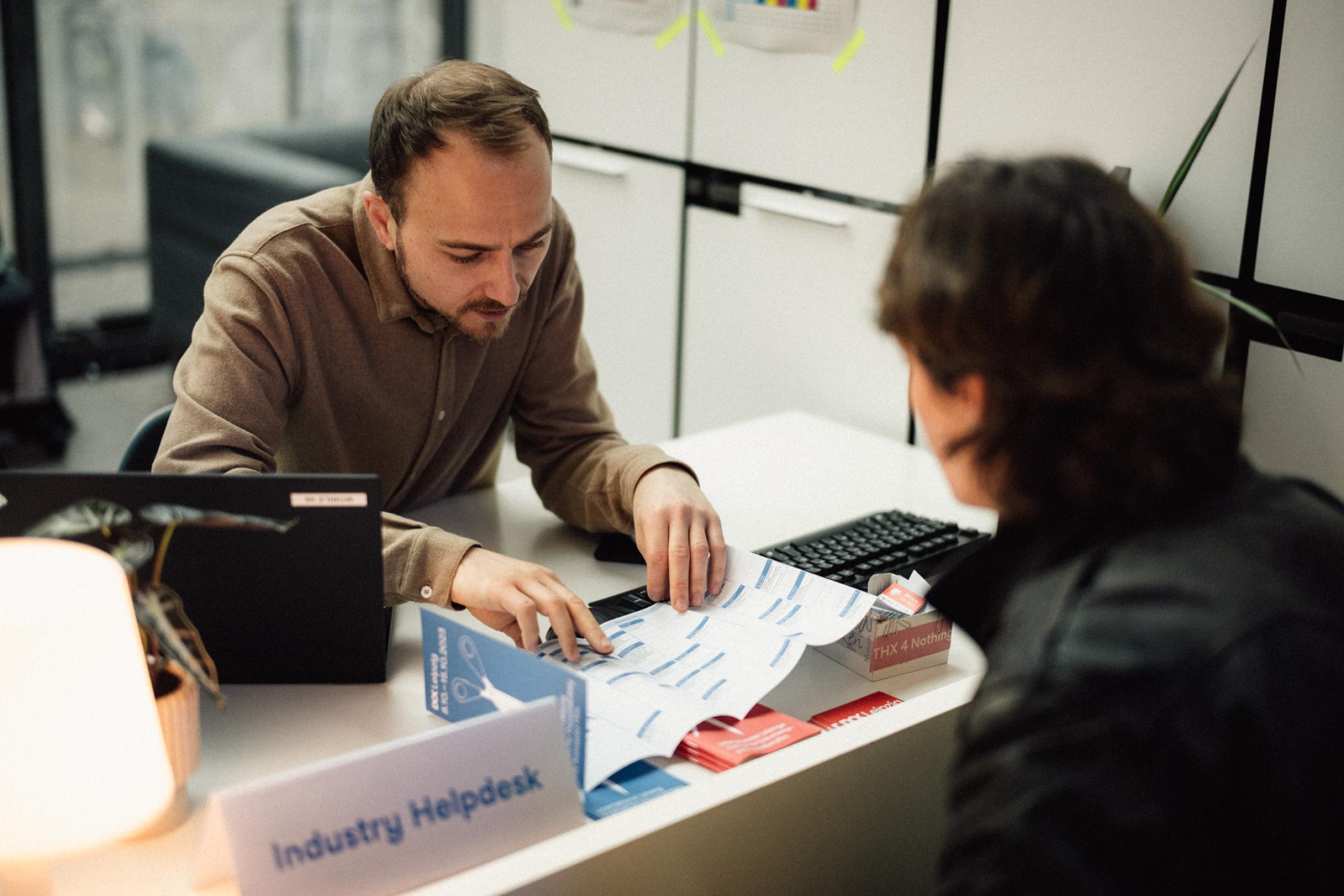 Two persons sit at a desk, looking at a leporello. On the desk a sign with "Industry Helpdesk".