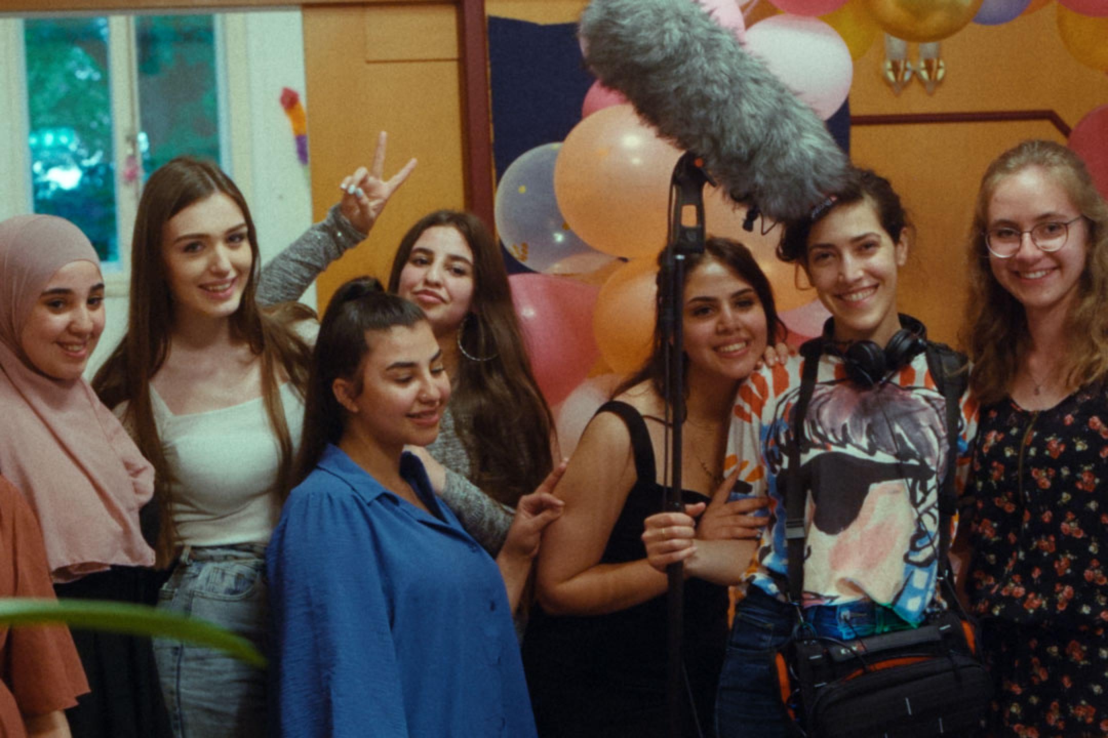 A group of seven young women pose for a picture. One of them holds a boom pole with a huge microphone.