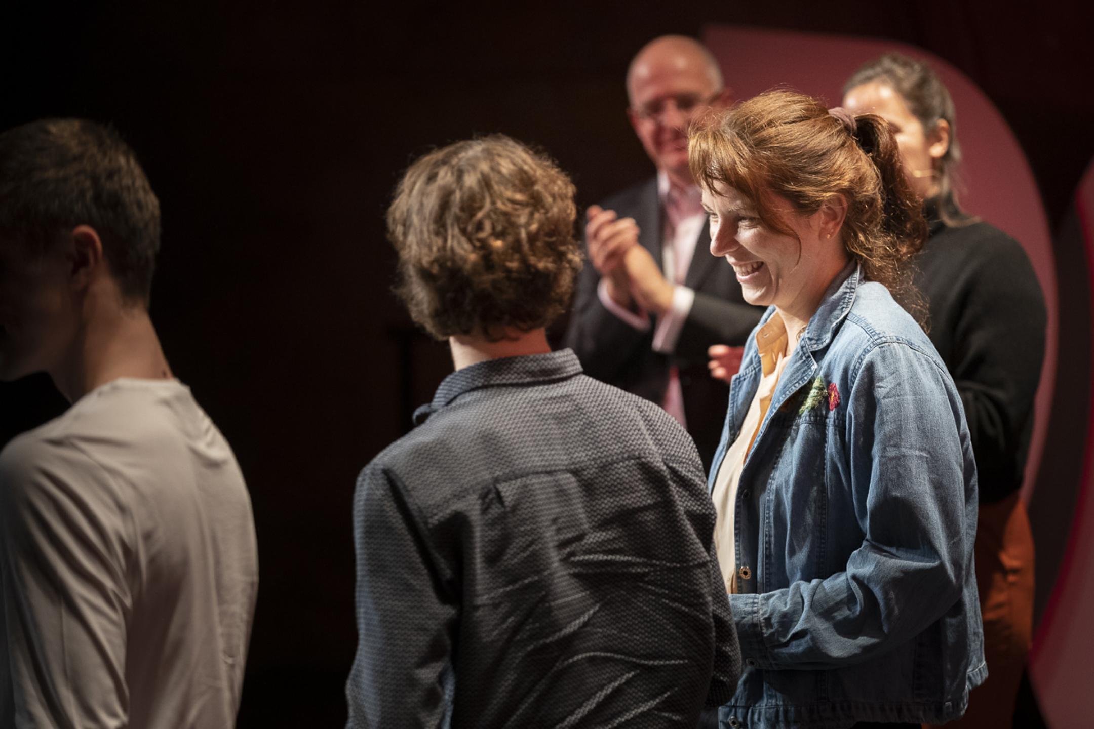 Fünf Personen stehen im Rampenlicht auf einer Bühne. Eine Person applaudiert, die Person in der Mitte strahlt.