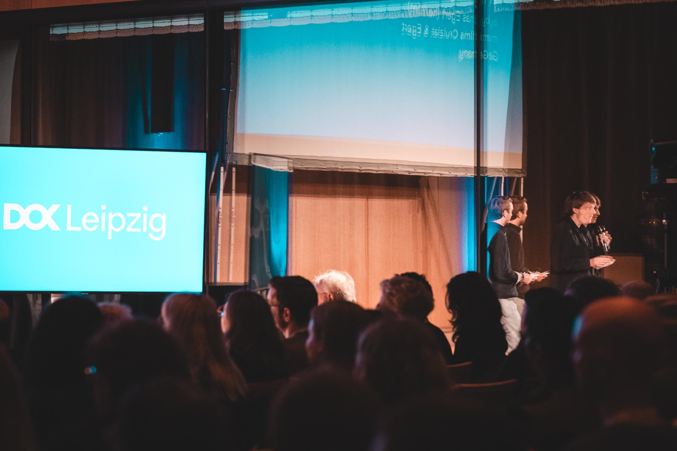 A presentation room viewed through the reflections of the windows. Two persons stand with hand microphones in front of an audience. Behind them a blue screen with the DOK Leipzig logo.