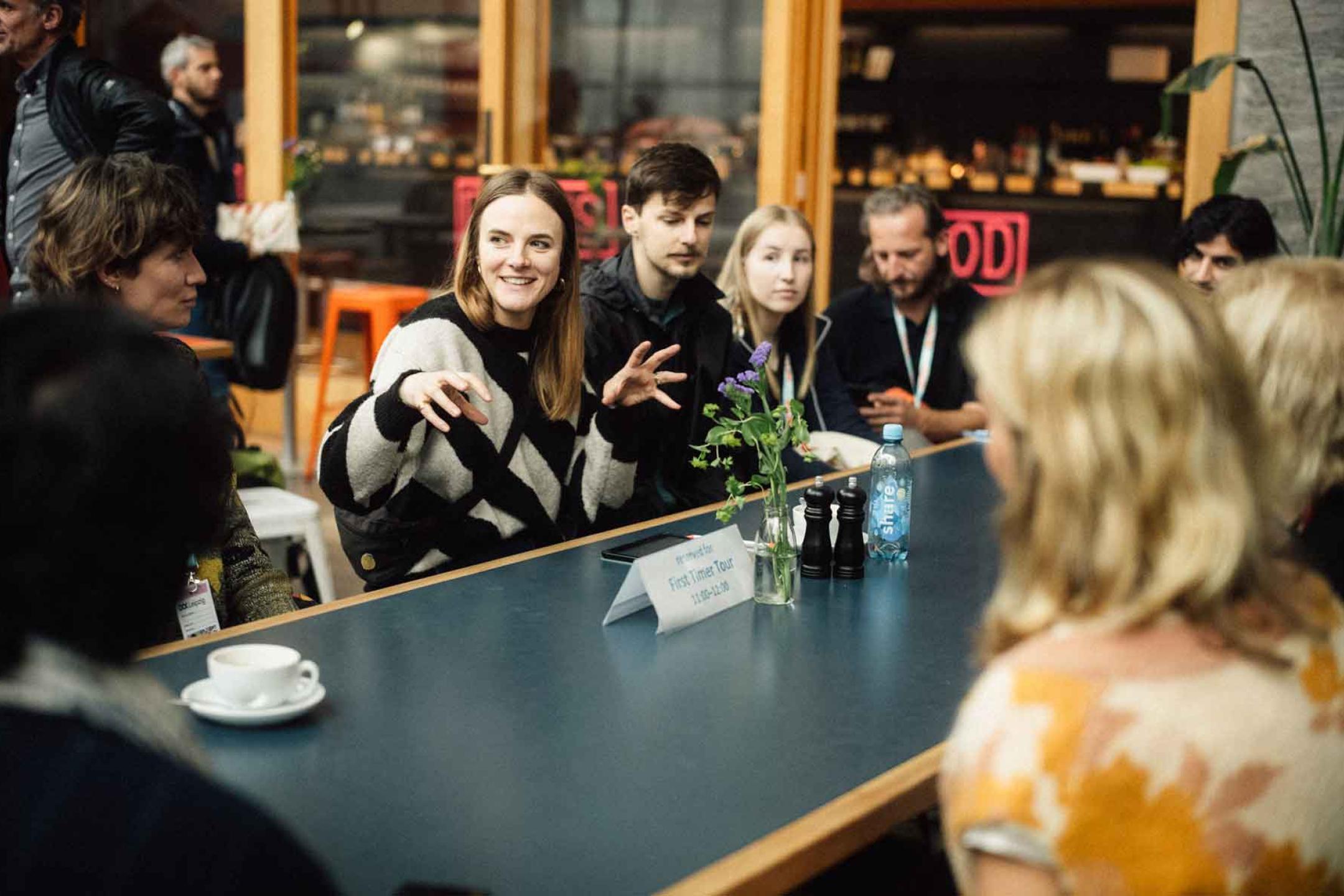 A group of people sit at a table listening to one woman in the circle talking animatedly
