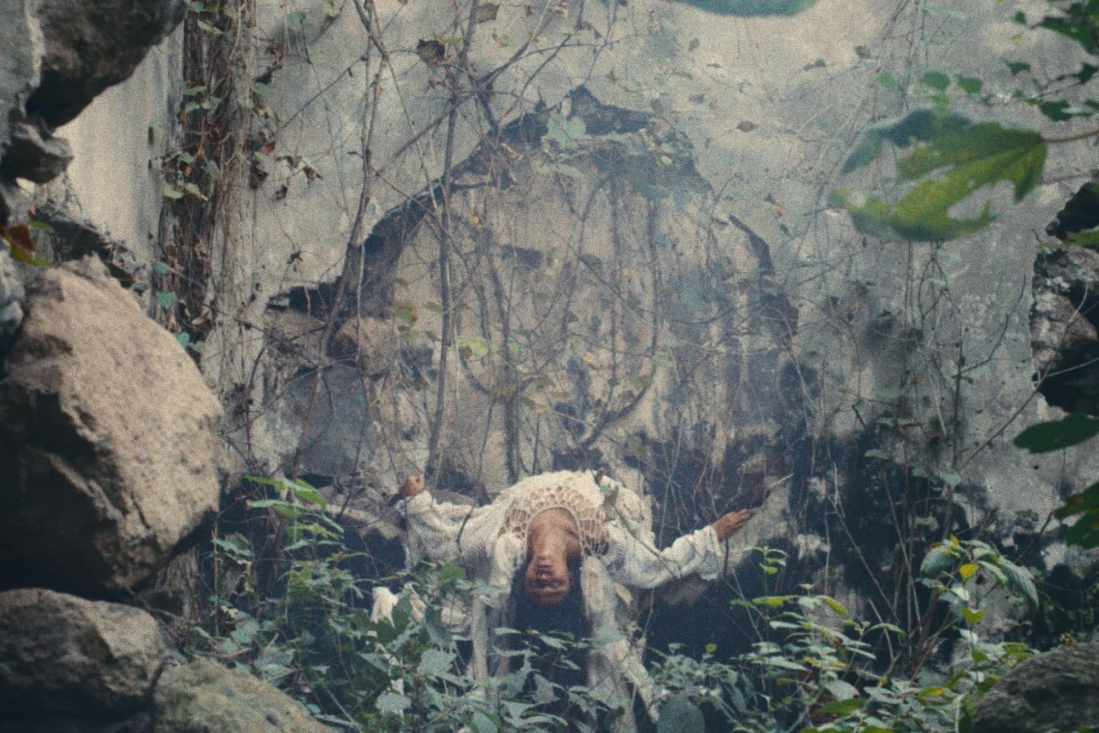 In a forest in front of a stone wall, a person is floating on her back with her head hanging down towards the camera