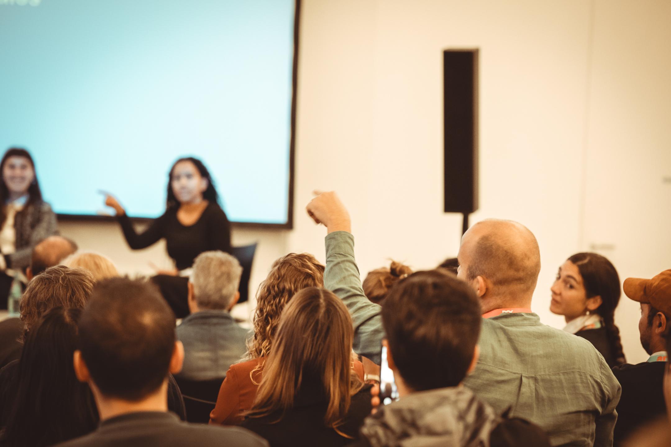 A person in the audience raises their hand and points one of the panelists on stage to ask them a questions