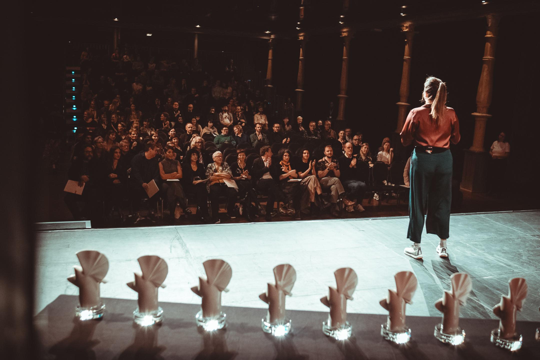 Der Schaubühne Saal voller Publikum, auf der Bühne steht eine Moderatorin und fein säuberlich sieben Goldene und Silberne Tauben