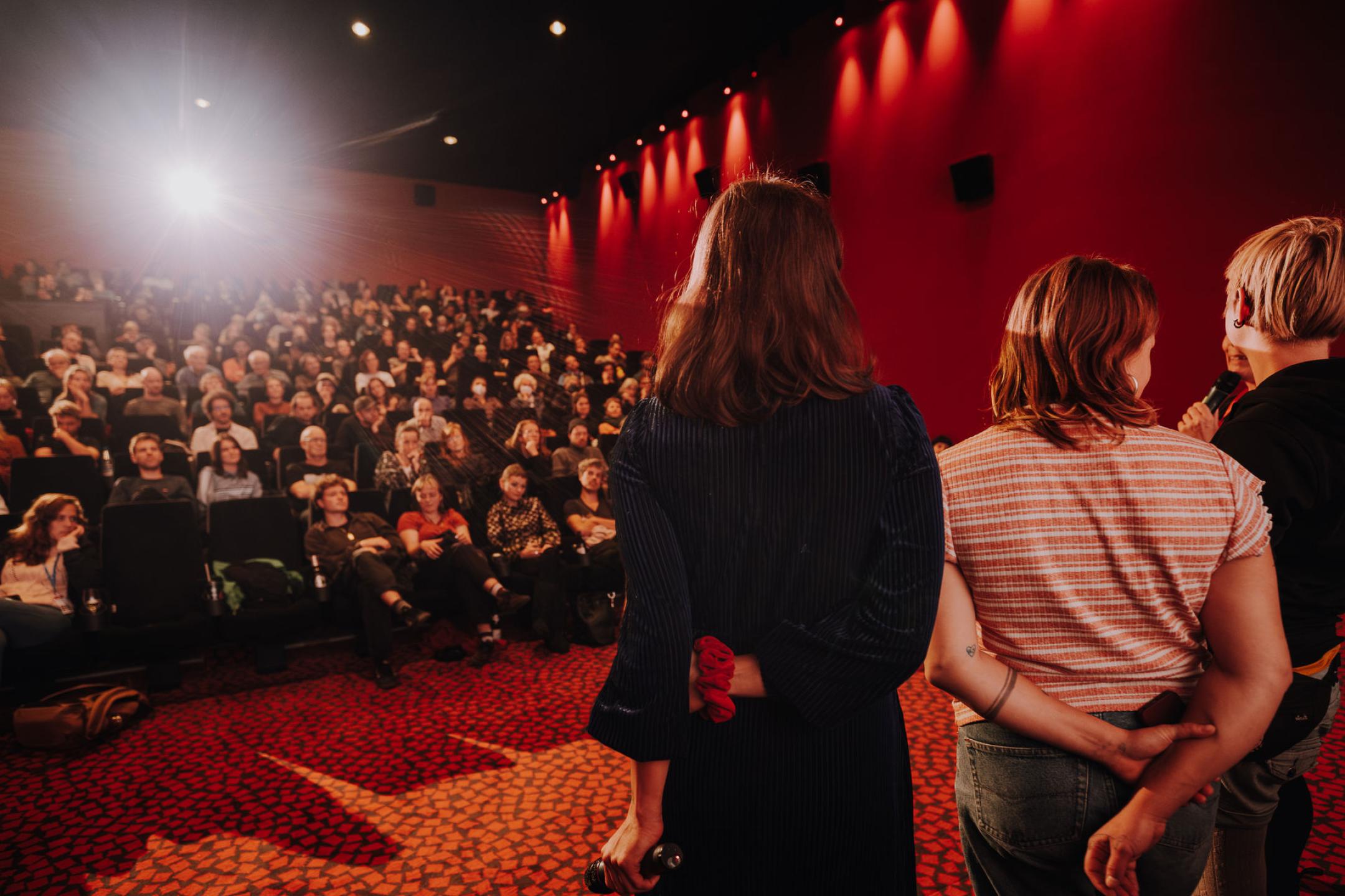 Im Kino stehen drei Menschen auf der Bühne vor einem vollen Saal im strahlenden Licht der Projektion