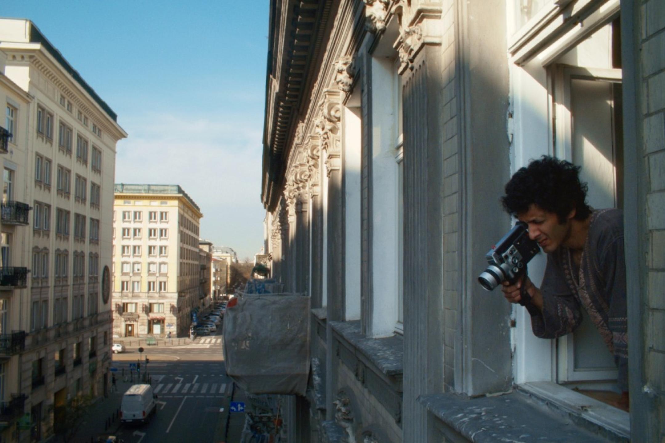 A curly haired man films with his video camera out of his window