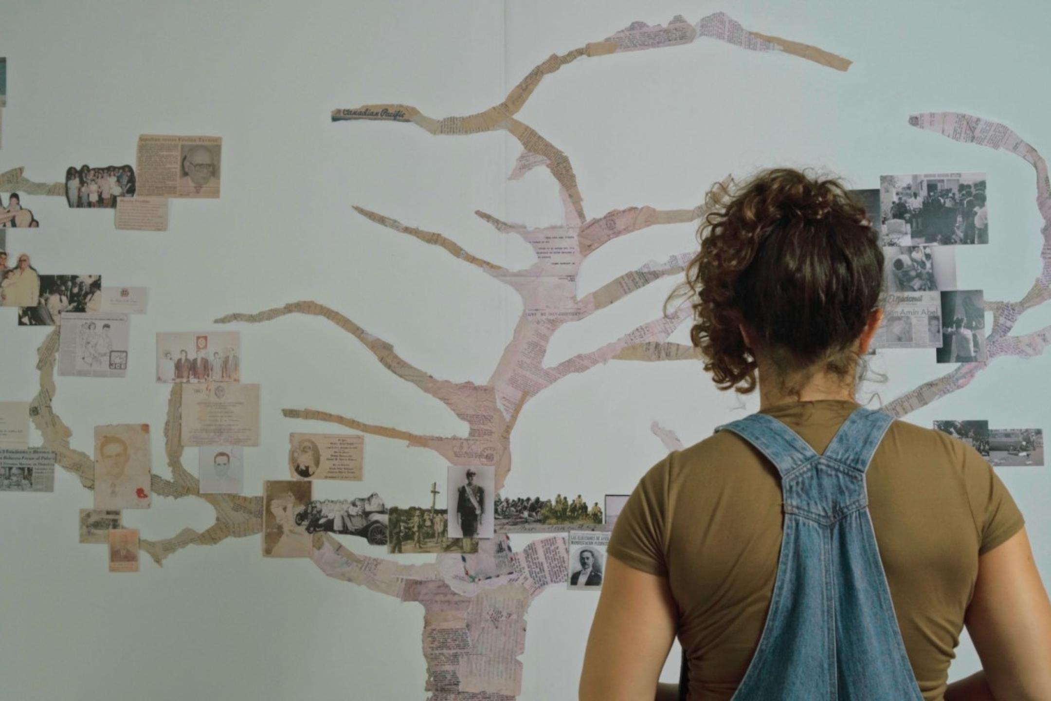 A person in jean dungarees stands in front of a family tree pained on a wall with many black-and-white photographs and documents