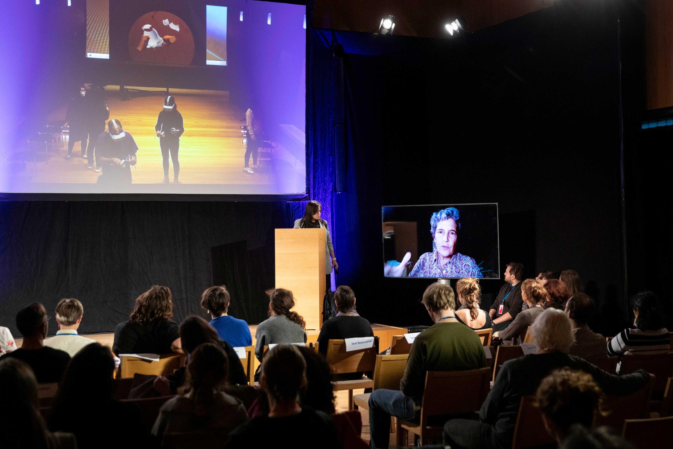 An audience watches a big screen on which people wearing VR glasses are walking around