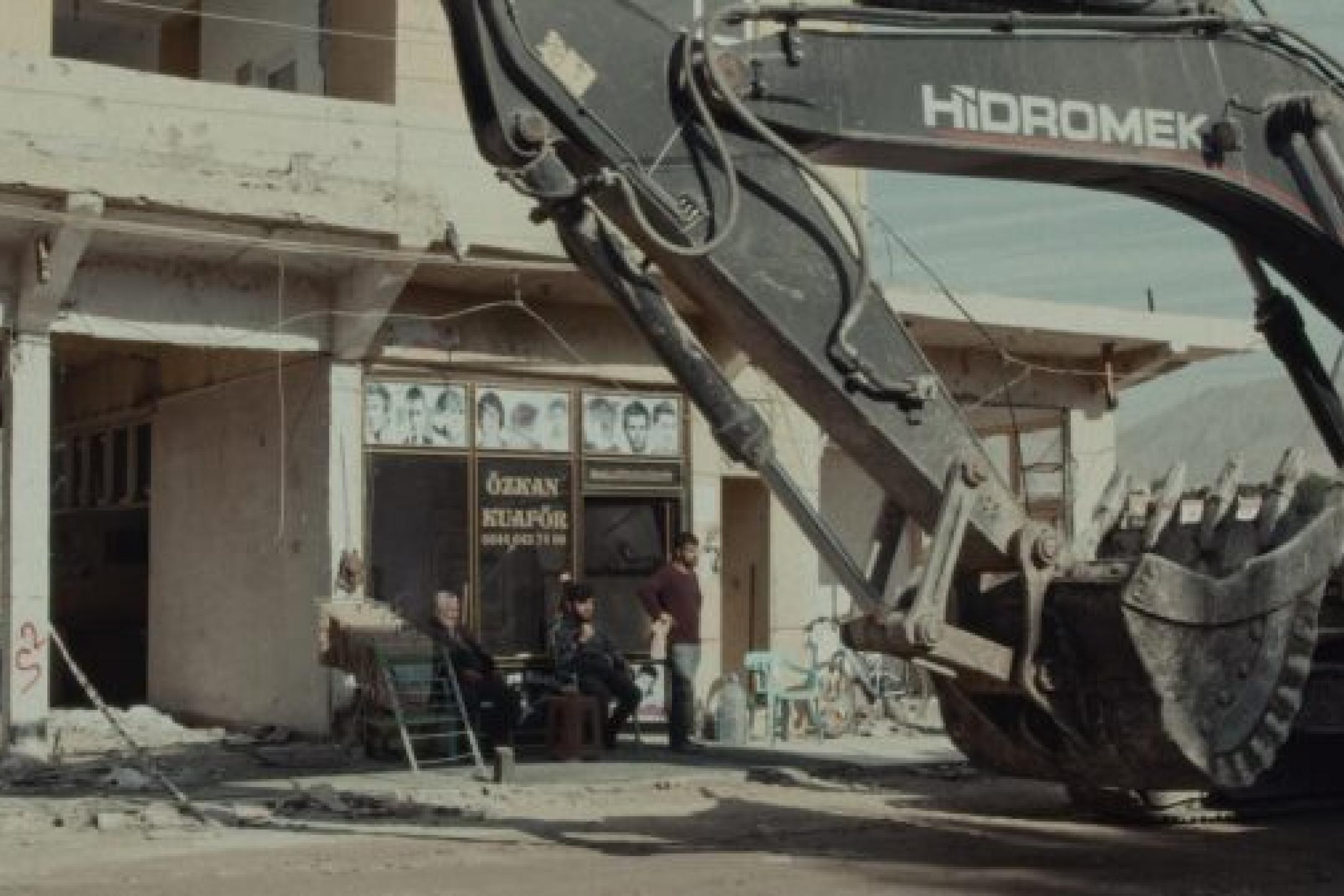 An excavator is parked in front of the storefront of a hair dresser. Several men sit in the front of the store and critically look over to the machine