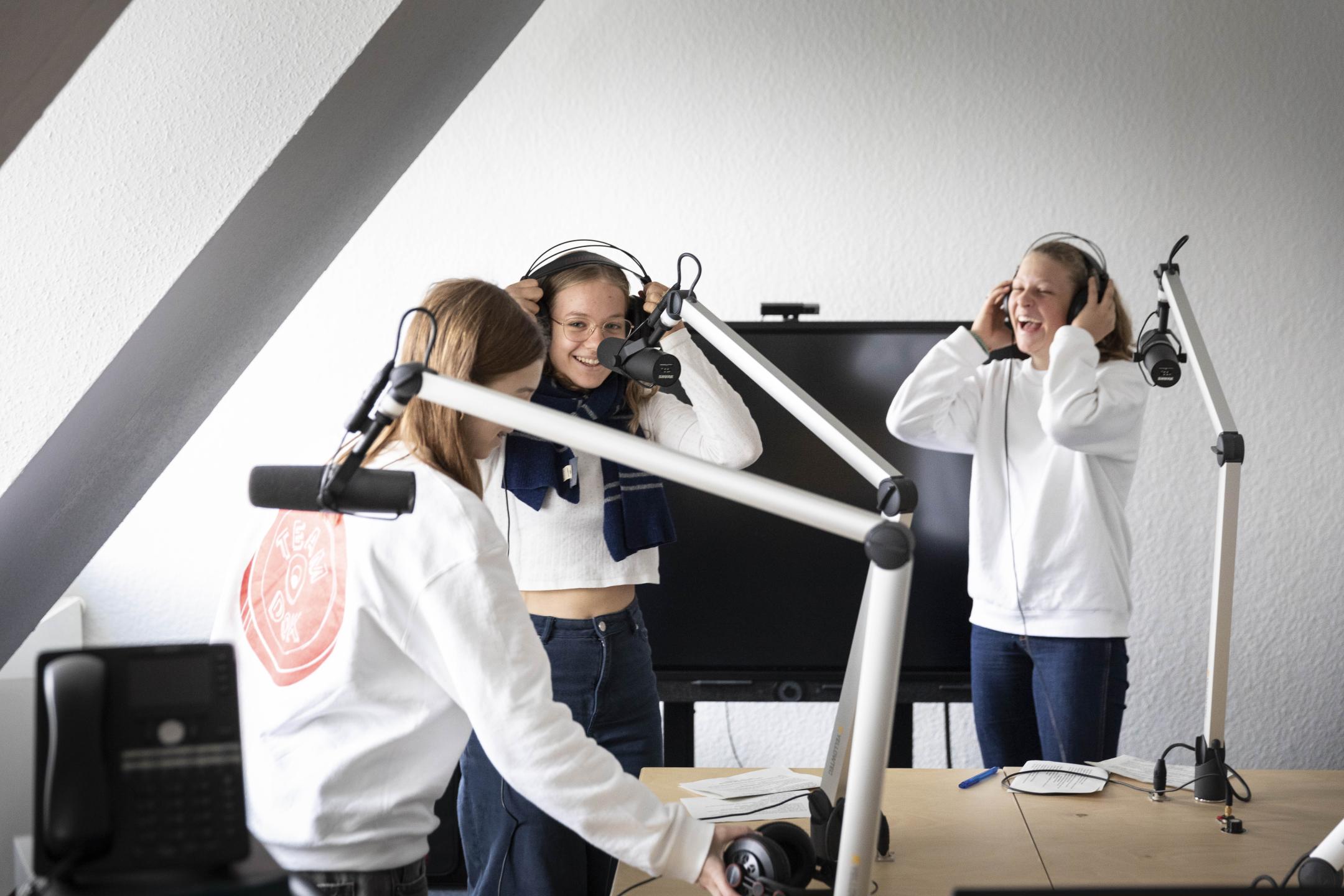 Drei Jugendliche in DOK Spotter Pullovern und mit Kopfhörern stehen um Radiomikrophone herum und lachen miteinander