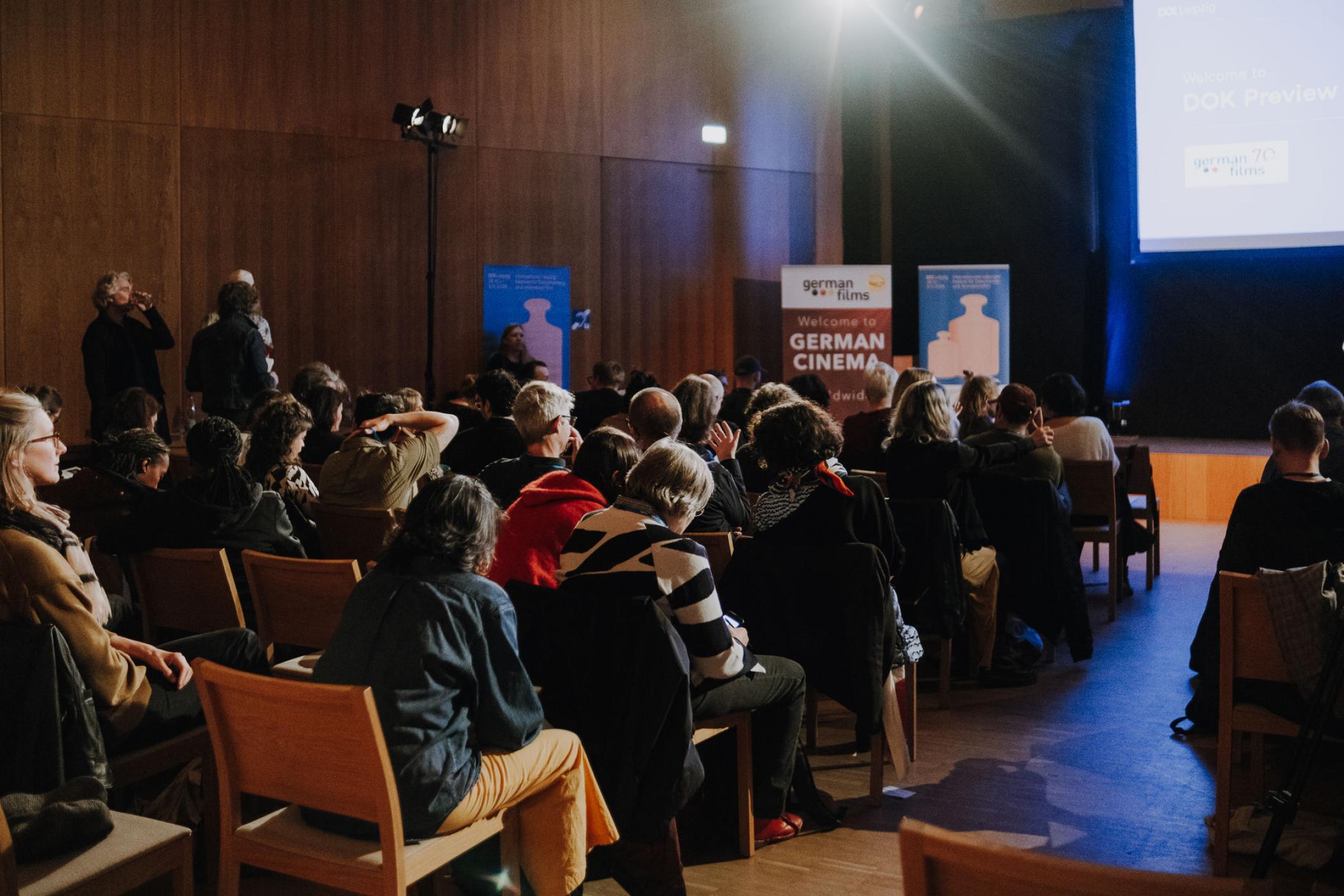 An audience is waiting for the show to start, next to the stage is a German Films banner and a Dok Leipzig poster