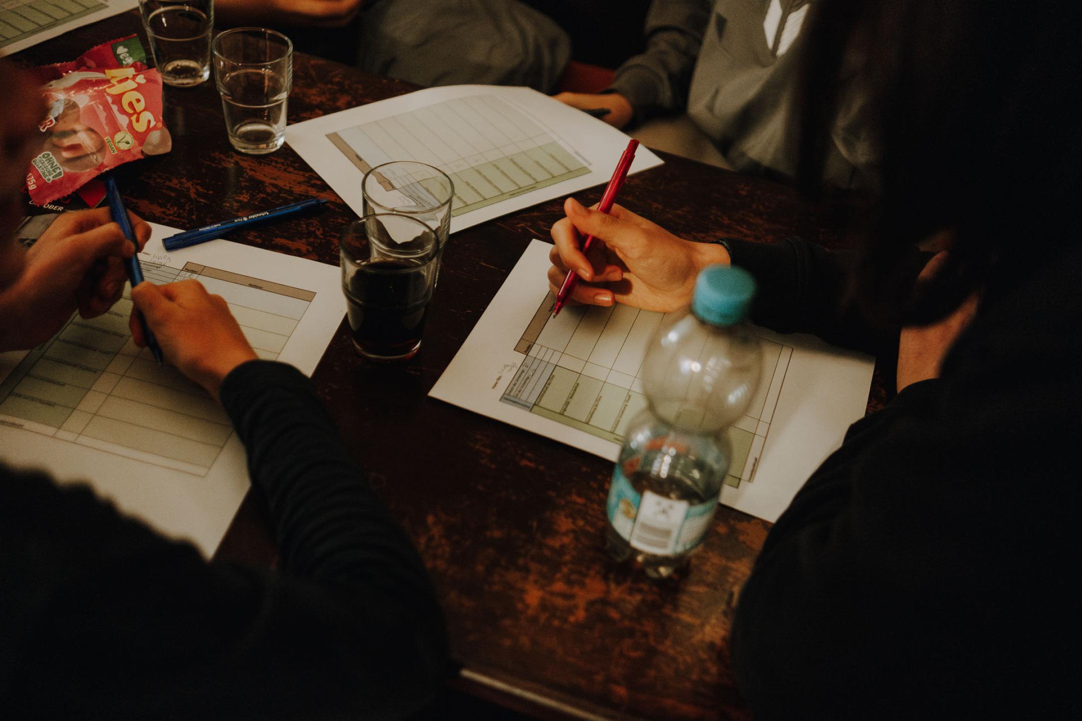 Hände von jungen Menschen auf füllen auf einem Tisch voller Snacks und Getränke eine Liste aus