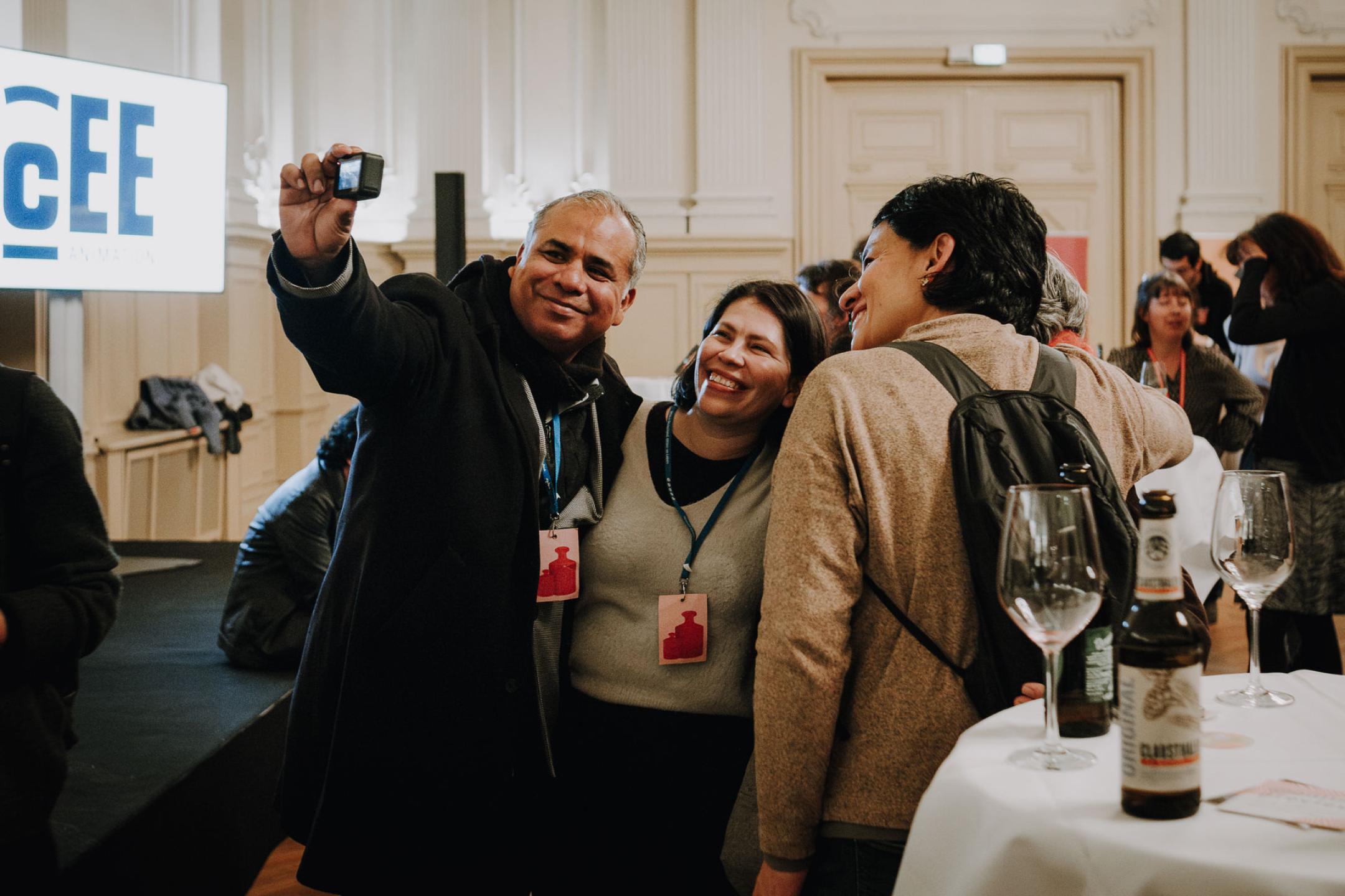 In a crowded room, three filmmakers are taking a group picture of themselves with a tiny camera