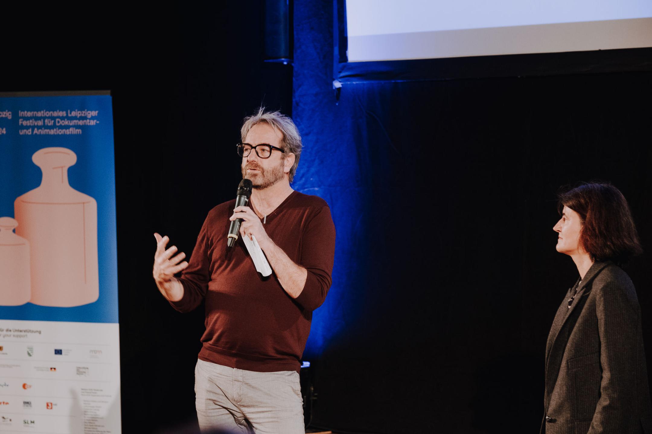 On stage, next to a Dok industry banner, a person is speaking into a microphone and gesticulates with his right arm